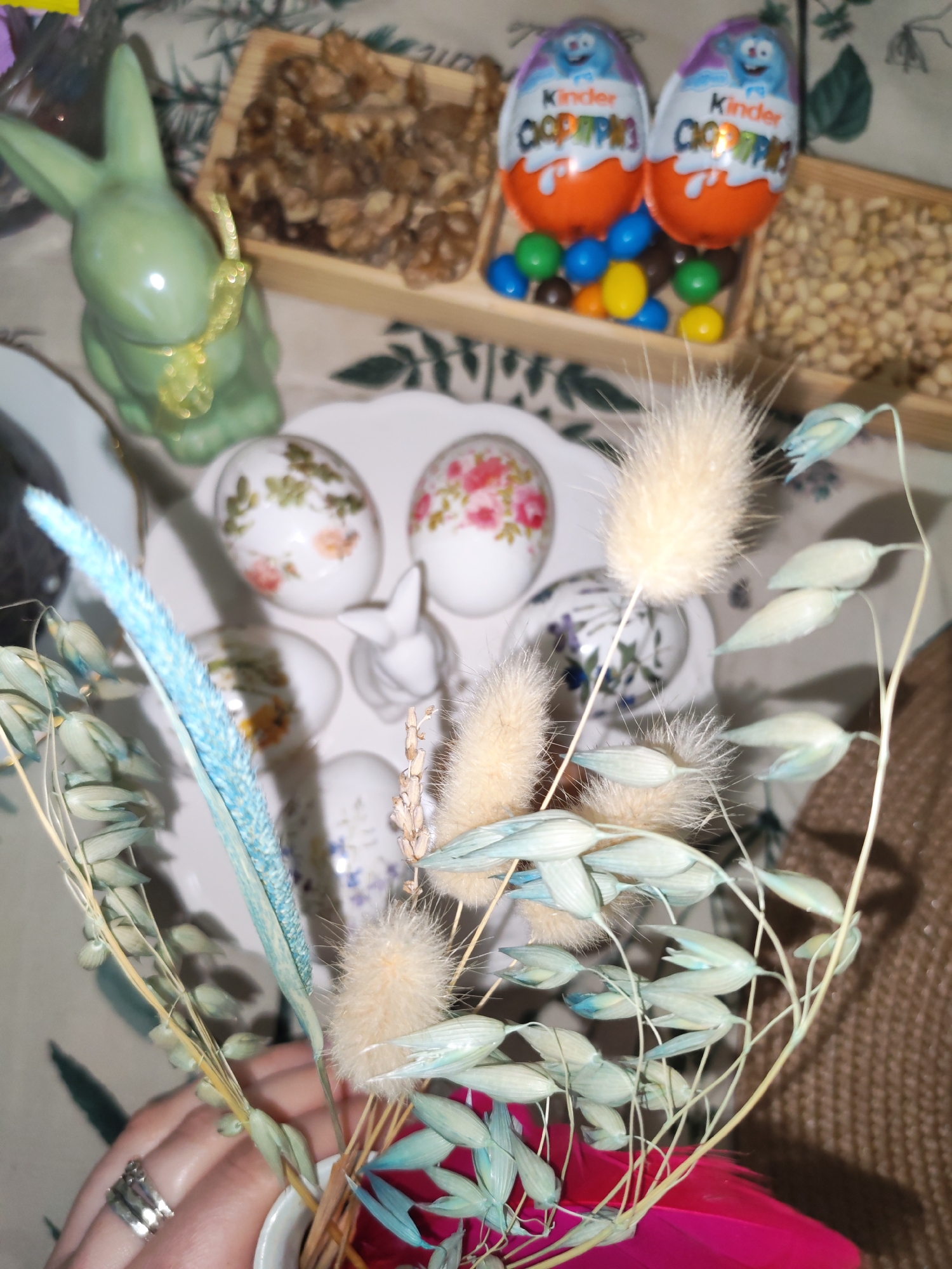 The scene is an indoor setting, possibly a home. The focus is on a close-up display of Easter-themed items.
Objects present include:
- A green ceramic bunny figurine with a yellow ribbon.
- A white plate holding decorated eggs with floral patterns, and a small white ceramic bunny figurine.
- Two Kinder Surprise chocolate eggs, surrounded by multi-colored candies.
- Two wooden trays: one filled with walnuts, and another with peanuts.
- A bouquet of dried flowers in a vase. The flowers have been dyed in shades of blue and beige.
- A hand is partially visible, wearing rings.
The background shows a tablecloth with a floral pattern.
The location is not discernible from the image content.
There are no people present, only a hand holding a bouquet.
There is no activity.
There are no clear interactions.
The environment is festive and related to Easter.
The location cannot be inferred to be in Izhevsk, Russia.