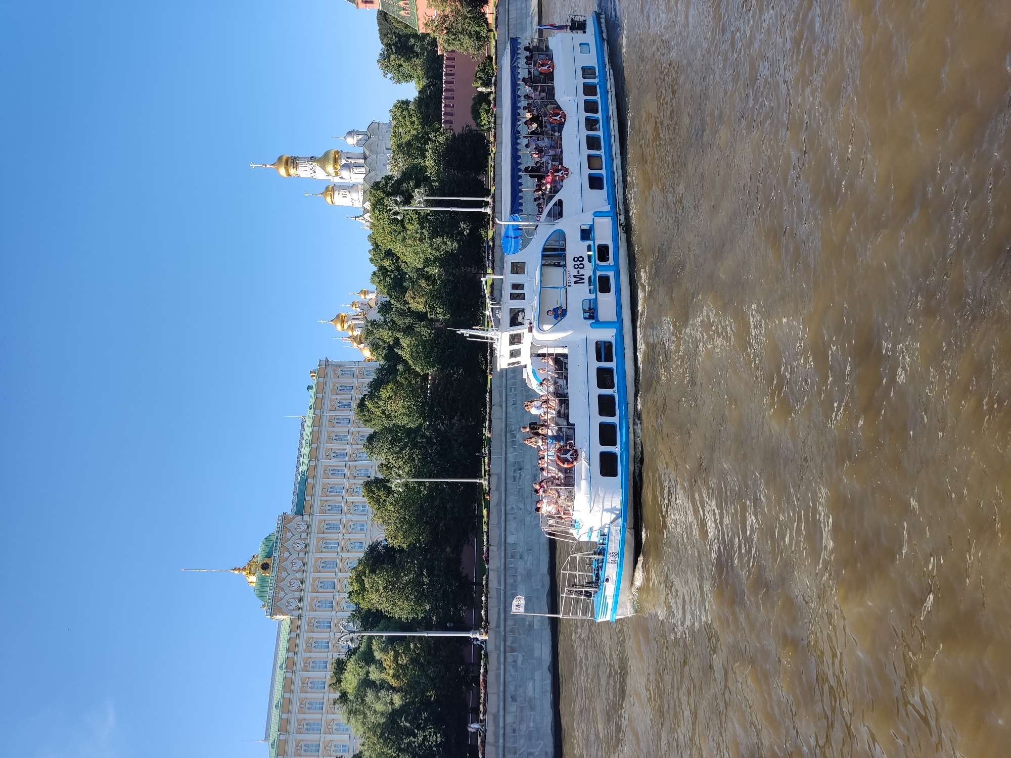 The image depicts a bright, sunny day with a clear blue sky over a city. In the foreground, a river with murky brown water flows from left to right. A white and blue tour boat, identified as "M-88," is sailing on the river, carrying numerous passengers on its upper deck, who are mostly looking out at the scenery. The boat has a covered seating area and a flag with "M-88" on it.
Behind the river is a stone embankment, followed by a lush green line of trees. Beyond the trees, a grand architectural complex is visible. This complex includes a large, pale yellow building with a green roof and a golden spire, suggestive of a palace or official building. To the right of this structure, several white churches with golden domes stand out against the sky. Further to the right, a red brick wall, likely part of a fortress, is partially visible. The overall scene suggests a major tourist attraction in a city with significant historical and architectural landmarks.