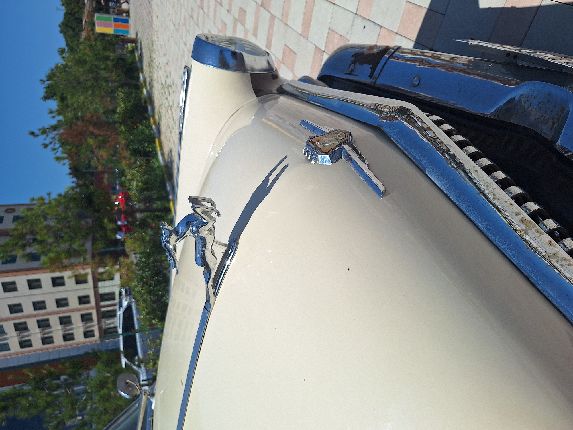 An exterior shot captures the front section of a vintage, cream-colored automobile, possibly parked outdoors in an urban setting. The hood is adorned with a chrome hood ornament depicting a leaping gazelle. A chrome trim piece runs along the hood's edge, and a distinctive emblem is mounted on the front fender. The car's front headlight, encased in chrome, is visible on the right. The vehicle's chrome bumper, exhibiting some signs of wear and rust, is prominent in the foreground, and the grille is partially visible beneath it.
The background reveals a tiled pedestrian area. Beyond this, a modern, multi-story building with numerous windows stands out. Green trees are interspersed throughout the scene, and a bright blue sky dominates the upper portion. In the far distance, figures of people and other vehicles can be vaguely discerned. The presence of a colorful art installation, featuring a grid of colored squares, adds a touch of vibrancy to the background. The lighting suggests a sunny day. The context of the city of Khudat, Azerbaijan, is provided, indicating the geographical setting of this scene.