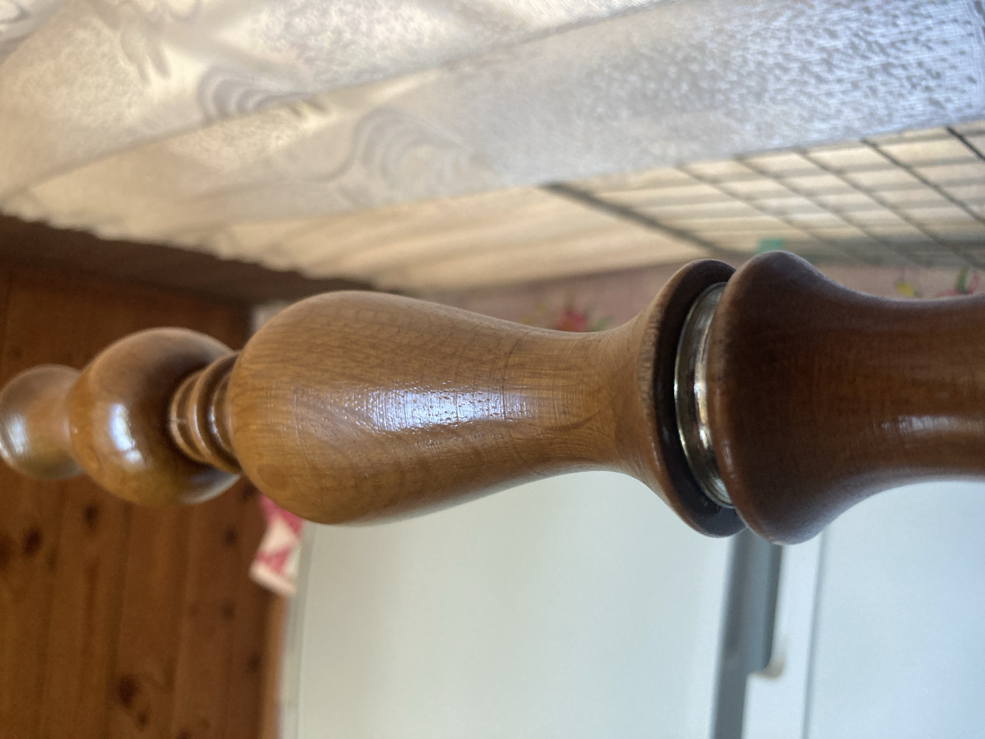 The image is a close-up, low-angle shot of a wooden decorative finial, likely part of a piece of furniture or architecture. The finial is made of polished, light brown wood with a glossy finish, showing turned details and a bulbous shape. It is positioned vertically, with the camera focused on the mid-section. In the background, the scene is blurred, showing hints of a wooden ceiling, a white surface which could be a wall or another piece of furniture, and a white lace curtain. There are no people or animals present in the image, nor any indication of specific activities or interactions. The environment appears to be indoors, in what could be a domestic setting. No specific location within the city of Ракиты, Russia can be determined from the image.