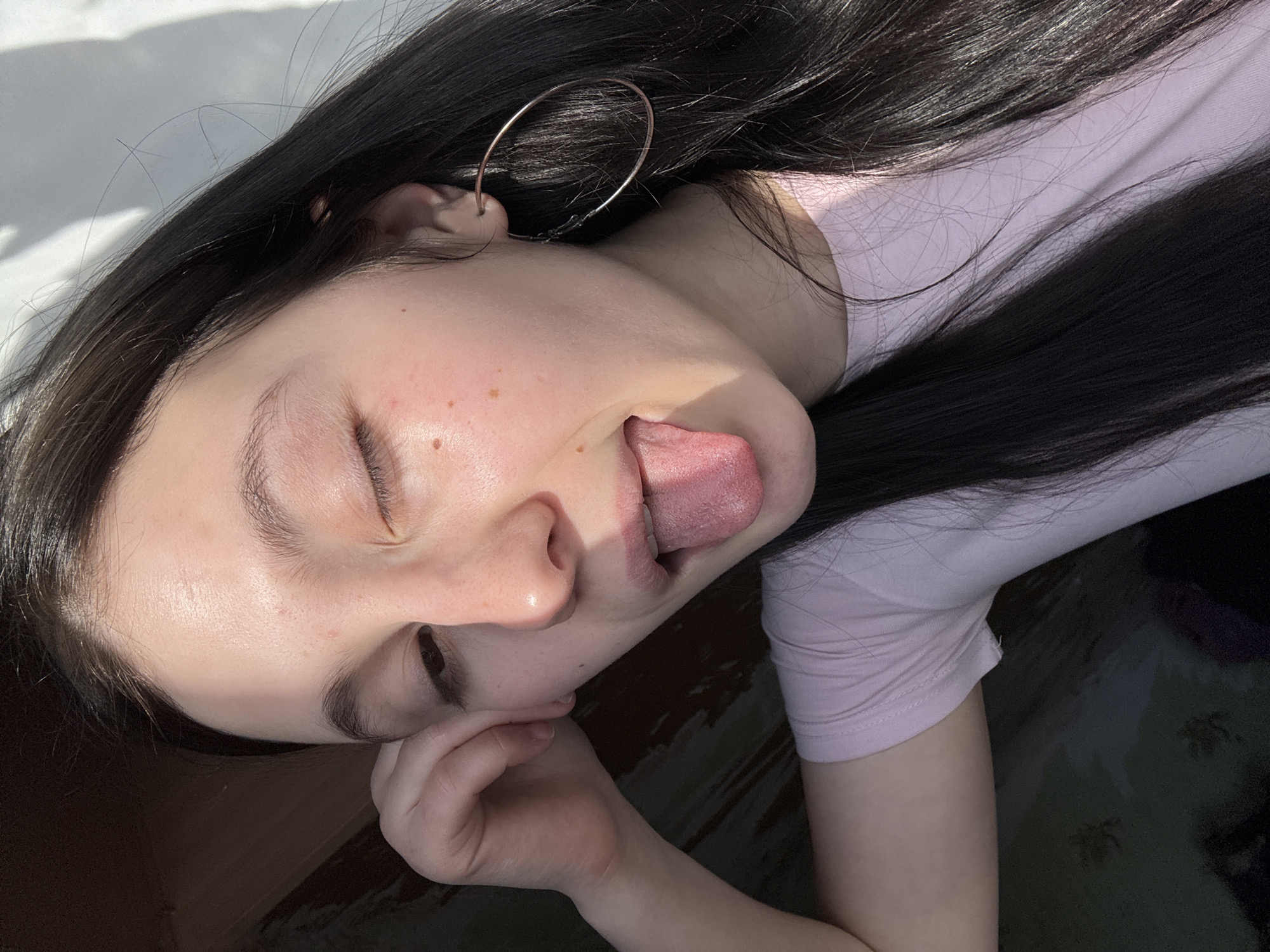 A young woman with long, dark hair and a large hoop earring is lying on her side, possibly outdoors. Her eyes are closed and she is sticking her tongue out. She is wearing a light-colored shirt. Her hand is near her face. The location is not discernible from the image.