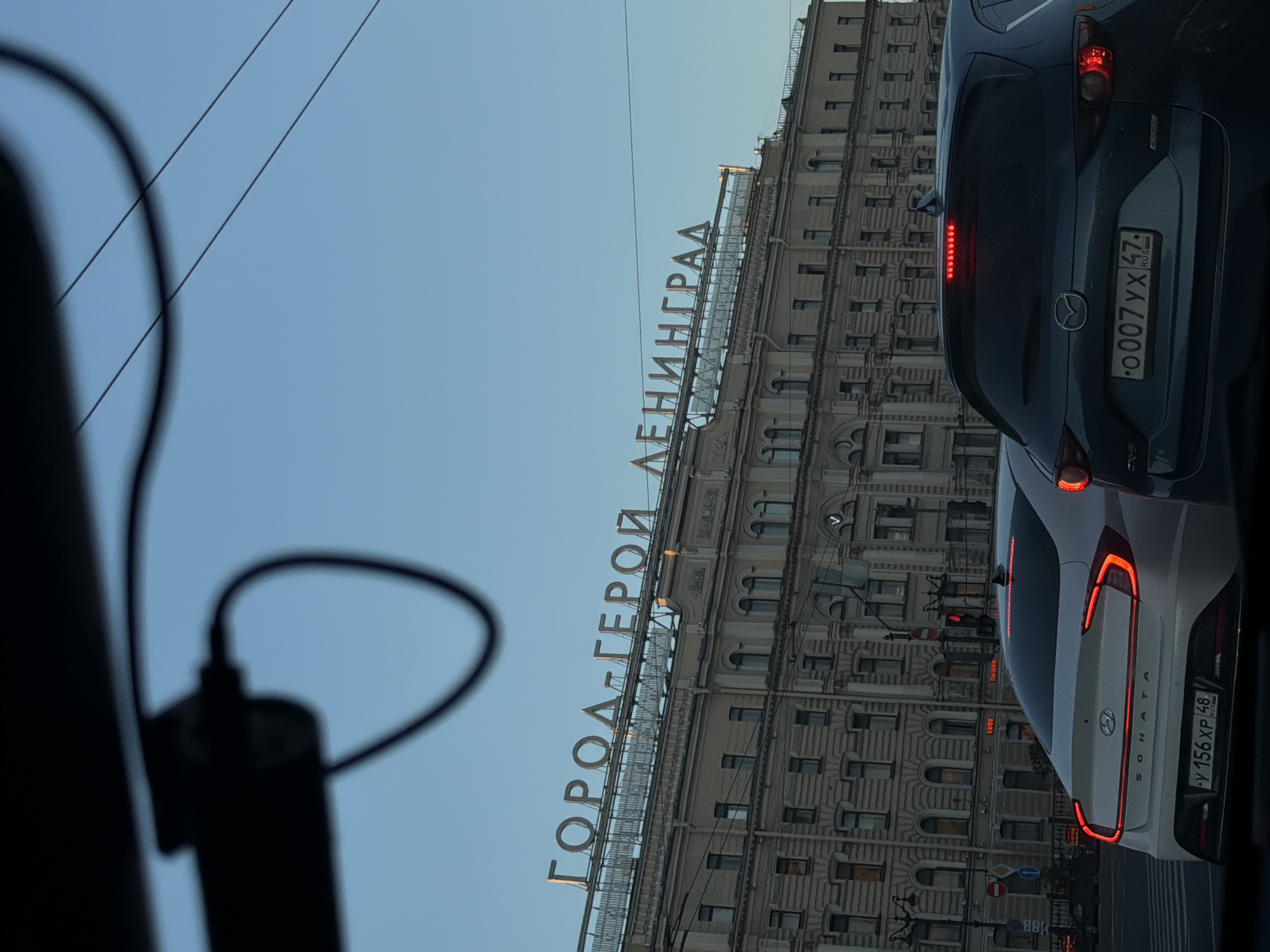 The image shows a street scene in Saint Petersburg, Russia, with a large building in the background adorned with a prominent sign that reads "ГОРОД-ГЕРОЙ ЛЕНИНГРАД" (Hero City Leningrad). The building is an example of classical architecture, with multiple stories, numerous windows, and decorative elements. In the foreground, two cars are visible, a white Hyundai Sonata and a dark blue Mazda CX-5, stopped at a traffic light. The Hyundai Sonata has its brake lights illuminated and a license plate that reads "У 156 ХР 48". The Mazda CX-5 also has its brake lights on and a license plate that reads "О007УХ 47 RUS". The sky is clear and blue. The overall scene captures a typical urban environment in Saint Petersburg.