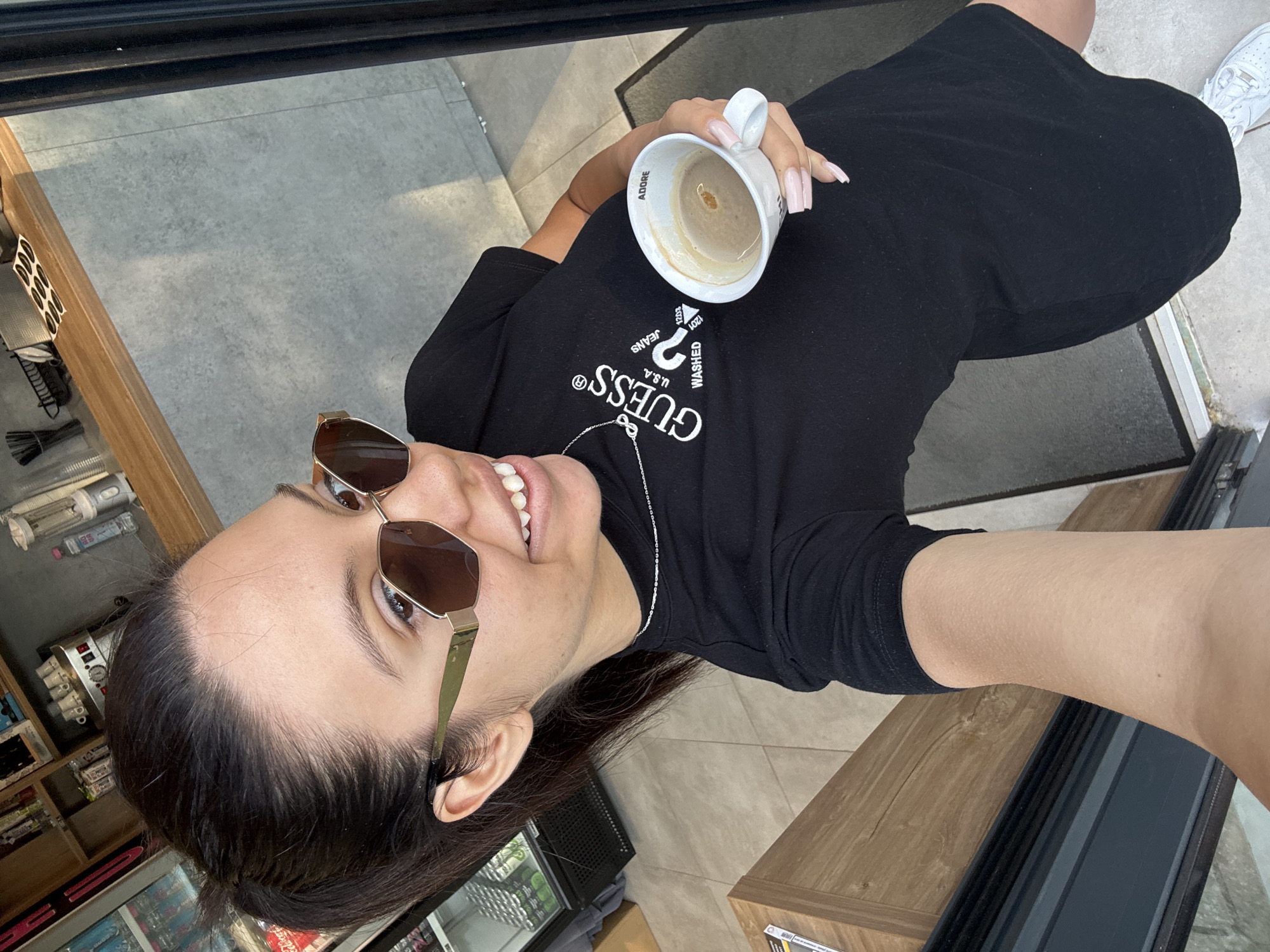 A young woman with dark hair pulled back in a ponytail and wearing large, gold-rimmed sunglasses takes a selfie while holding a white coffee mug in her right hand. She is wearing a black t-shirt with the "GUESS" logo printed on it, and a thin silver necklace. The woman's left arm is bent, and her hand is resting on her leg. The mug in her right hand is partially full of coffee and has "ADORE" written on it. She has long, light pink acrylic nails.
The background appears to be an indoor cafe or shop. To the left, there is a refrigerated display case with shelves and products inside. Behind her, there is a wooden counter with various items, including cups and a blender. The walls are concrete, and the floor is tiled. The overall lighting suggests it is daytime, possibly sunny, as indicated by shadows. The woman is standing in a doorway or open area, with a black metal pillar to her right. The environment suggests a commercial setting in Sofia, Bulgaria.