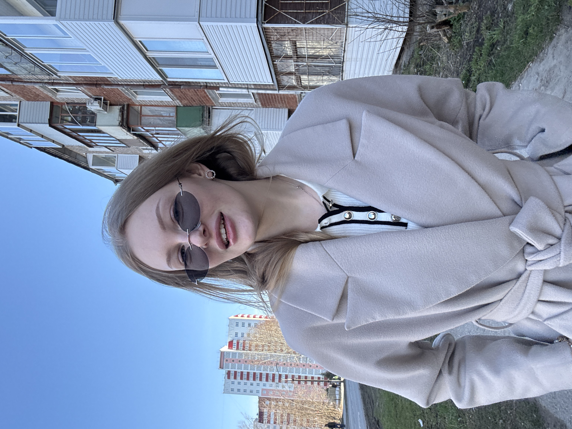 A young, fair-skinned woman with blonde hair, wearing sunglasses and a beige coat, is the primary subject of this outdoor photograph. She is smiling slightly at the camera.
Behind the woman are apartment buildings, typical of urban residential areas. Based on the architectural style, the location is likely in the city of Perm, Russia.
The environment includes a concrete sidewalk or walkway. There are also patches of grass and bare trees, suggesting the photo was taken in early spring or late fall. The sky is visible in the background, appearing blue.
The woman appears to be standing still, possibly posing for the photograph. There are some other people in the far background but no interaction can be identified.