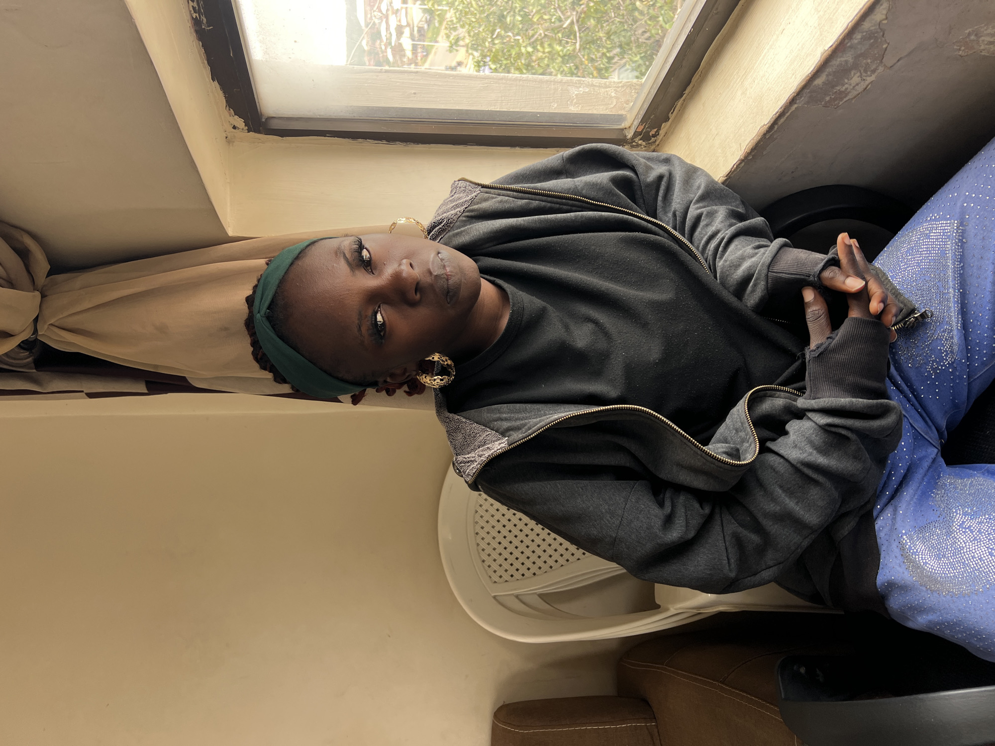 A medium shot captures a young Black woman seated in a plastic chair by a window, looking directly at the camera. She is wearing a dark grey zip-up hoodie over a black t-shirt, blue pants with silver embellishments, a green headband, and large gold hoop earrings. Her hands are clasped in her lap. The environment appears to be an interior space, with a beige wall to her left and a window with a view of an urban landscape in Jos, Nigeria, to her right. The window frame is aged and shows signs of wear. A portion of a brown couch is visible to the left of the chair. The lighting is natural, coming from the window.