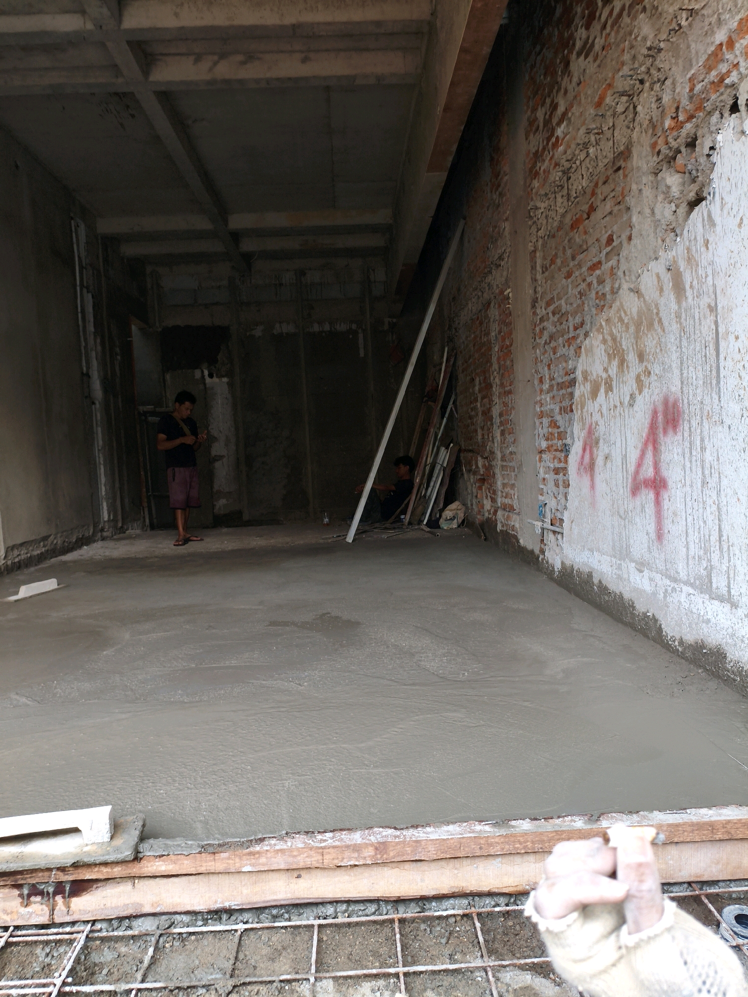 A medium-shot, eye-level, indoor photograph shows a construction site in Bekasi, Indonesia. In the foreground, rebar is visible beneath a wooden frame holding wet concrete, which has been freshly poured and smoothed. The floor of the room is covered in wet concrete. To the left, a white leveling tool rests on the edge of the concrete. A person stands in the middle-ground on the left, wearing a dark t-shirt and maroon shorts, facing away from the camera and looking down at their hands. Further back and to the right, another person is seated, partially obscured by construction materials including wooden planks and metal pipes. The walls of the room are unfinished brick and concrete. The wall on the right is textured and painted white with red markings that appear to be numbers or symbols. Overhead, wooden beams support a concrete ceiling.