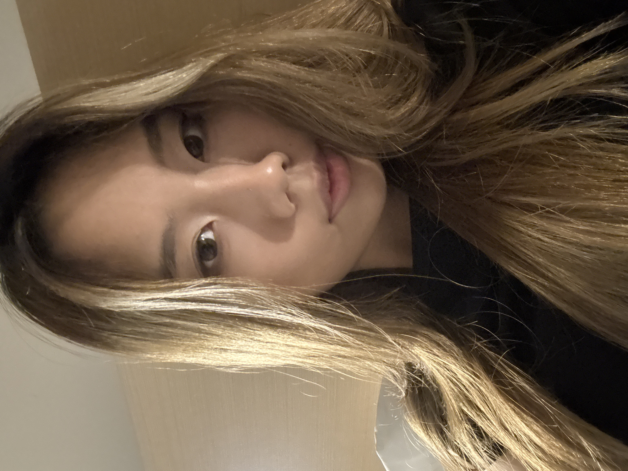 An extreme close-up selfie shows a young Asian woman with light brown hair and fair skin. She is wearing a black top. Her hair is long and straight with some waves at the ends, and it frames her face, obscuring part of the background. The background appears to be a wall with a textured pattern, possibly wood paneling, in a light brown hue. The lighting is soft, creating a subtle shadow on the left side of her face. The image is composed from a high angle, looking down at the subject. The setting does not provide specific clues to a location in Yuzhong District, China.