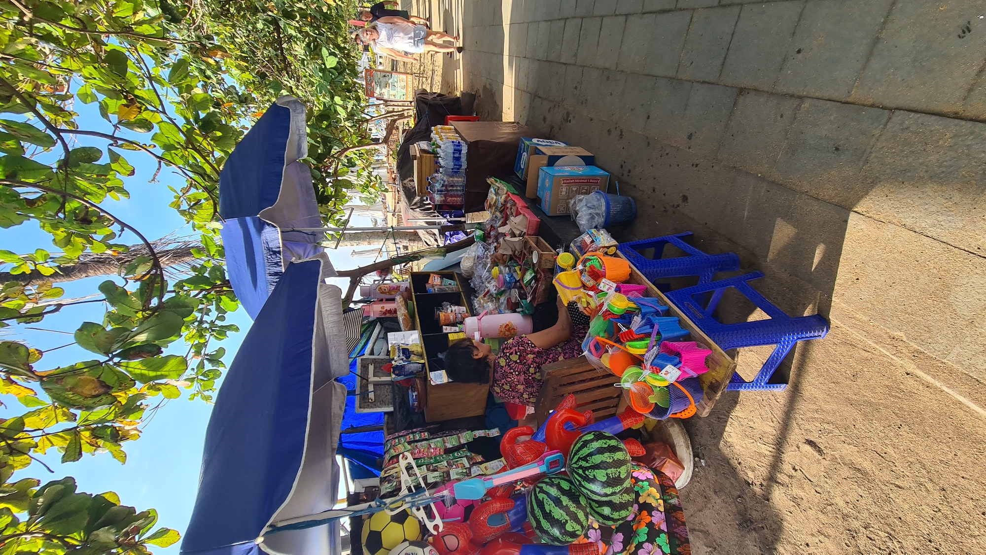 The image depicts a beachside market stall in Denpasar, Indonesia, likely situated along the popular tourist area of Sanur. The stall is under two blue and white striped awnings, shaded by lush green trees and a bright blue sky. The stall is filled with a variety of items, including inflatable toys like a yellow soccer ball and red Spiderman figures, as well as buckets and spades in various colors, suggesting it caters to beachgoers and families.
A woman with dark hair, wearing a floral-patterned top, is seated behind a table laden with goods. The table displays items such as drinks, snacks in packaging, and more beach toys. Behind her, shelves are stocked with bottles and containers. The ground in front of the stall is sandy, with a paved walkway to the right where two people are walking. One is a woman in a white tank top and shorts, and the other is a man in a dark shirt. In the background, beach umbrellas and palm trees are visible, characteristic of Denpasar's coastal landscape. The overall scene captures a typical vibrant and bustling atmosphere of a local market near a tourist beach.