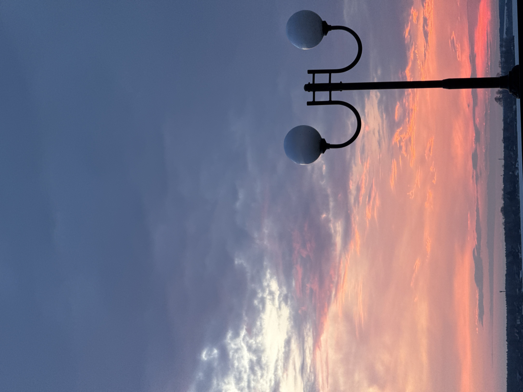 The image shows a lamppost with two globe lights against a dramatic sky with a mix of dark blue and shades of pink and orange, suggesting a sunset or sunrise. The lamppost is black, and its design features curved arms supporting each globe. The background sky has a gradient effect, transitioning from a darker, stormy-looking blue on the left to a vibrant display of sunset colors on the right. A line of trees and buildings is visible on the far right horizon. Given the prompt, the location is likely Votkinsk, Russia.