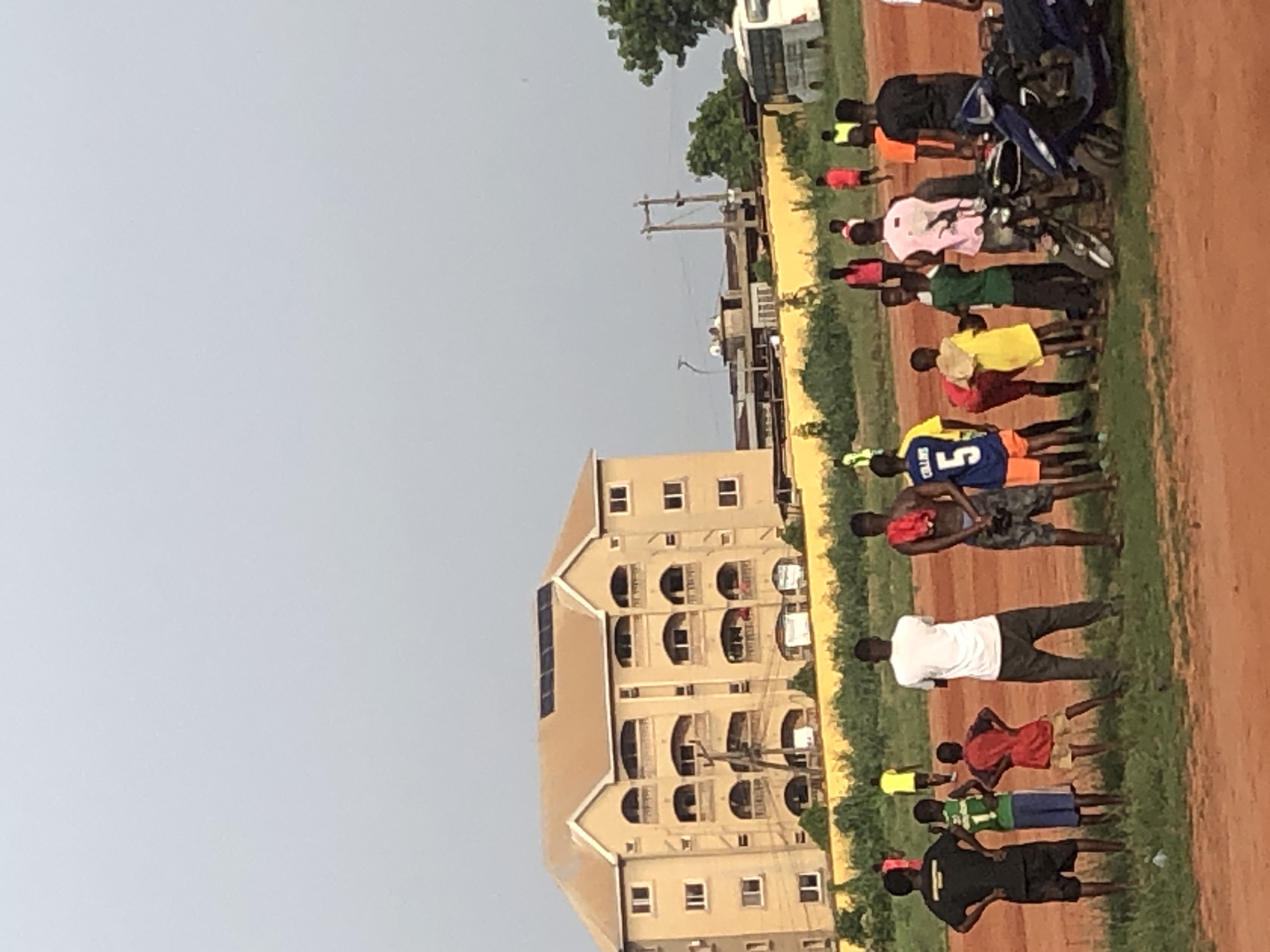 This outdoor image, taken in Ukwu-Abwa, Nigeria, shows a group of approximately 15-20 young men and boys gathered on a mixed dirt and grassy field under an overcast or hazy sky during daylight hours.
On the left, a multi-story building with a light-colored facade features several windows and arched balconies on each of its visible floors. Solar panels are installed on the building's roof. In the mid-ground, utility poles with power lines are visible.
The individuals are dressed in casual and athletic wear, including t-shirts and shorts, with some wearing colorful jerseys. Colors seen include red, green, yellow, blue, black, white, pink, and orange. One blue jersey prominently displays the number "5" and the name "COLLINS" on the back. They are standing in various relaxed postures, some appearing to be stretching or observing, suggesting a gathering for a sporting activity, likely football (soccer). Two motorcycles are parked near some of the individuals on the right side of the field. The ground is a blend of reddish-brown dirt and green patches of grass. In the far background, sparse greenery and a light-colored vehicle can be seen.
The scene depicts a communal event or training session, characterized by the informal gathering of young people in a local field.