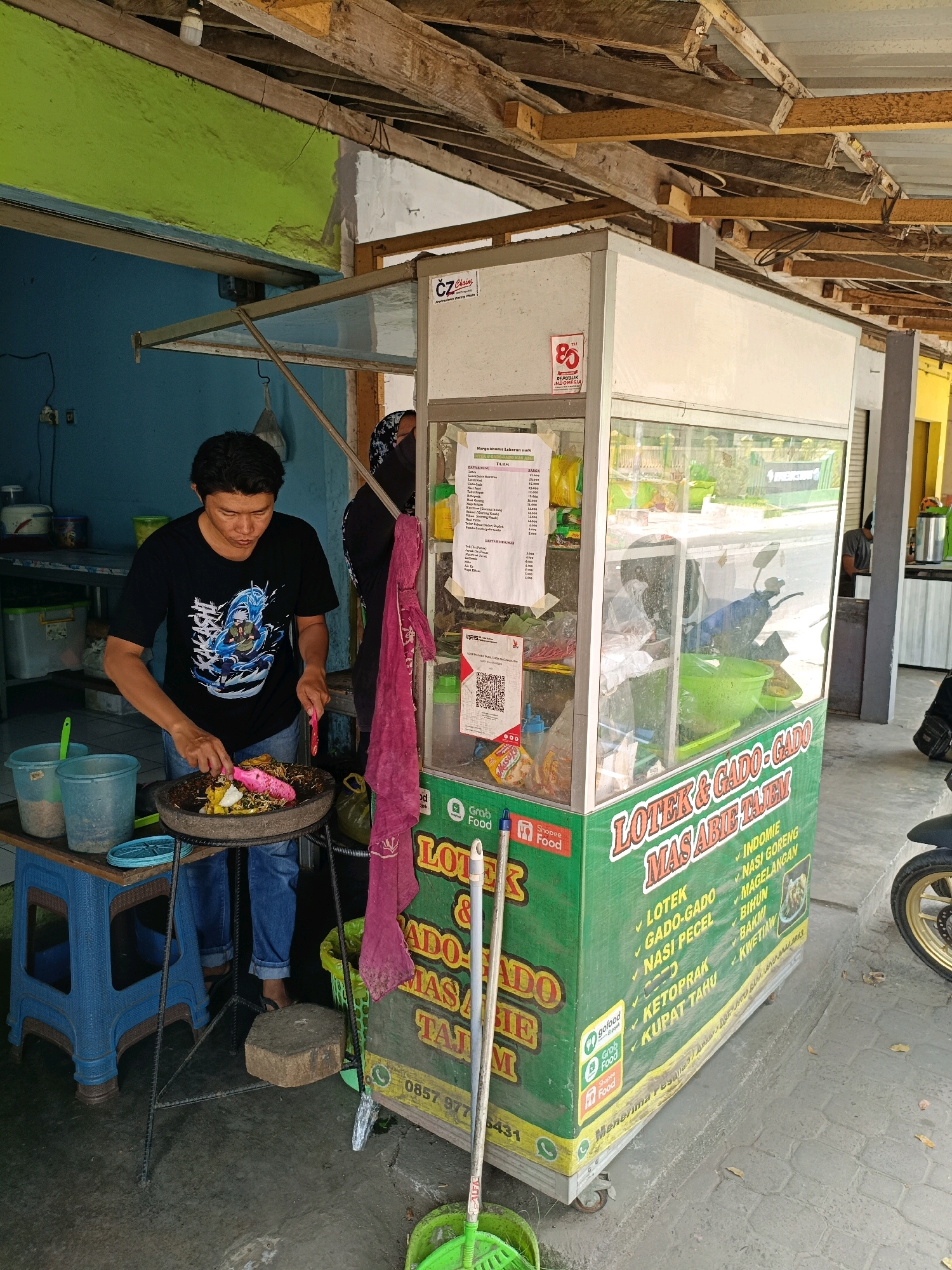 A street food stall with a man preparing food is pictured in Maguwoharjo, Indonesia. The stall is green and white, with the text "LOTEX & GADO-GADO MAS ABIE TAJEM" prominently displayed. A menu is visible inside the stall, along with a motorcycle. The man, wearing a black t-shirt and blue jeans, is actively engaged in preparing food in a mortar and pestle. He is holding a knife in his right hand and a utensil in his left, working over a stone mortar filled with ingredients. Two blue plastic buckets are placed to his left, one containing a green utensil and the other some grey material. A red cloth is draped over the stall's edge, near the man. The ground is paved with concrete. The stall is situated under a rustic wooden roof structure, suggesting an outdoor or semi-outdoor setting. The background shows a blue wall and what appears to be a kitchen area.