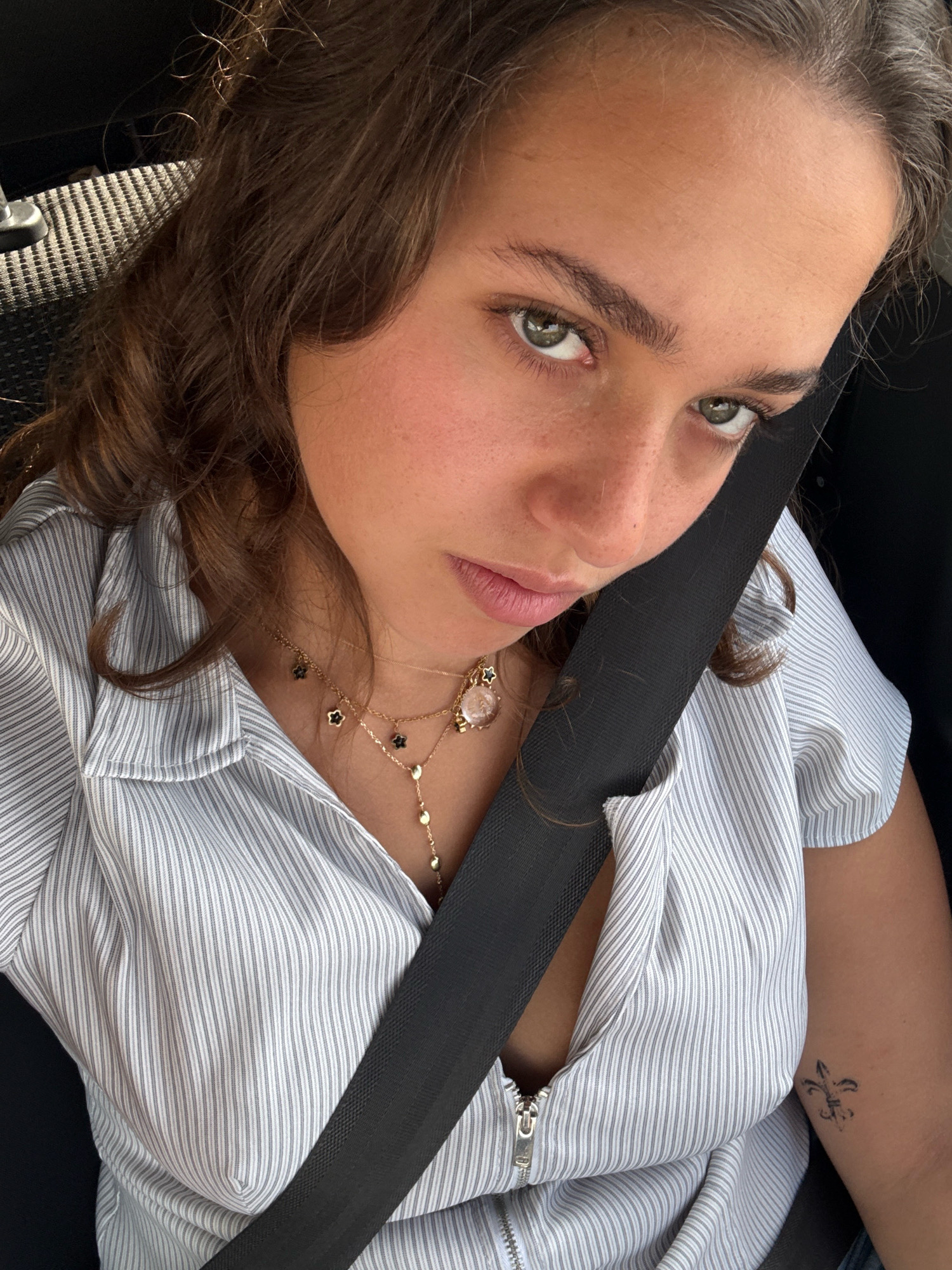 A young woman with long, brown, wavy hair and light green eyes is shown in a close-up, selfie-style photograph. She is wearing a light blue and white striped button-down shirt with a zipper partially undone, revealing a glimpse of her chest. She is also wearing a black seatbelt across her torso. Her right arm is visible, and a small tattoo of a fleur-de-lis is present on her upper arm. She has light makeup on, with emphasis on her eyes and lips. She is looking directly at the camera with a neutral expression. The background is dark, suggesting she is inside a vehicle. The photograph was taken in Busmantsi, Bulgaria.