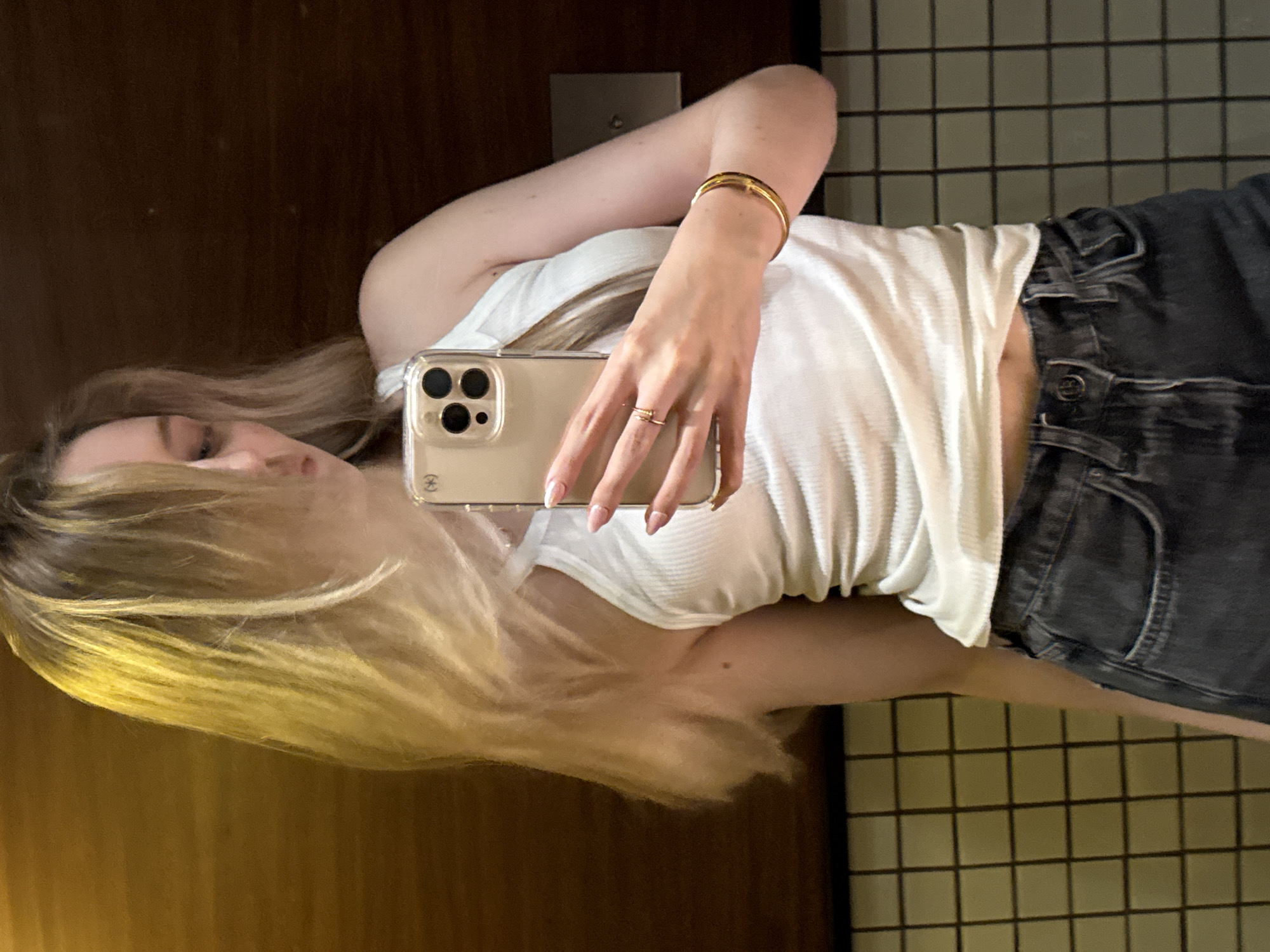 An indoor mirror selfie captures a young person, reflected horizontally in the image. She has long, flowing blonde hair and is wearing a ribbed white tank top paired with dark grey or black high-waisted jeans. A gold bracelet adorns her right wrist, and a small gold ring is visible on her left ring finger. Her fingernails are manicured in a french tip style.
She holds a light-colored smartphone, appearing to be a newer iPhone model with multiple cameras, encased in a clear cover. A small, partially obscured logo, likely an Apple logo, is visible on the phone's back. The visible portion of her face shows her right eye.
The background reflected in the mirror presents an indoor setting. To the person's left, a dark wooden wall or door is visible, featuring a small, rectangular metallic fixture. To her right, a wall covered in small, light-colored square tiles with dark grout lines is prominent, with a dark grid-like structure in the upper right corner. This suggests a modern interior space, potentially within a hotel or residence in Mexico City, Mexico.
Faintly discernible on the button of her jeans is a small, embossed brand name, possibly "G-Star RAW." The lighting is consistent with an indoor environment, and no specific time of day or weather can be inferred. The image portrays a casual act of self-documentation.