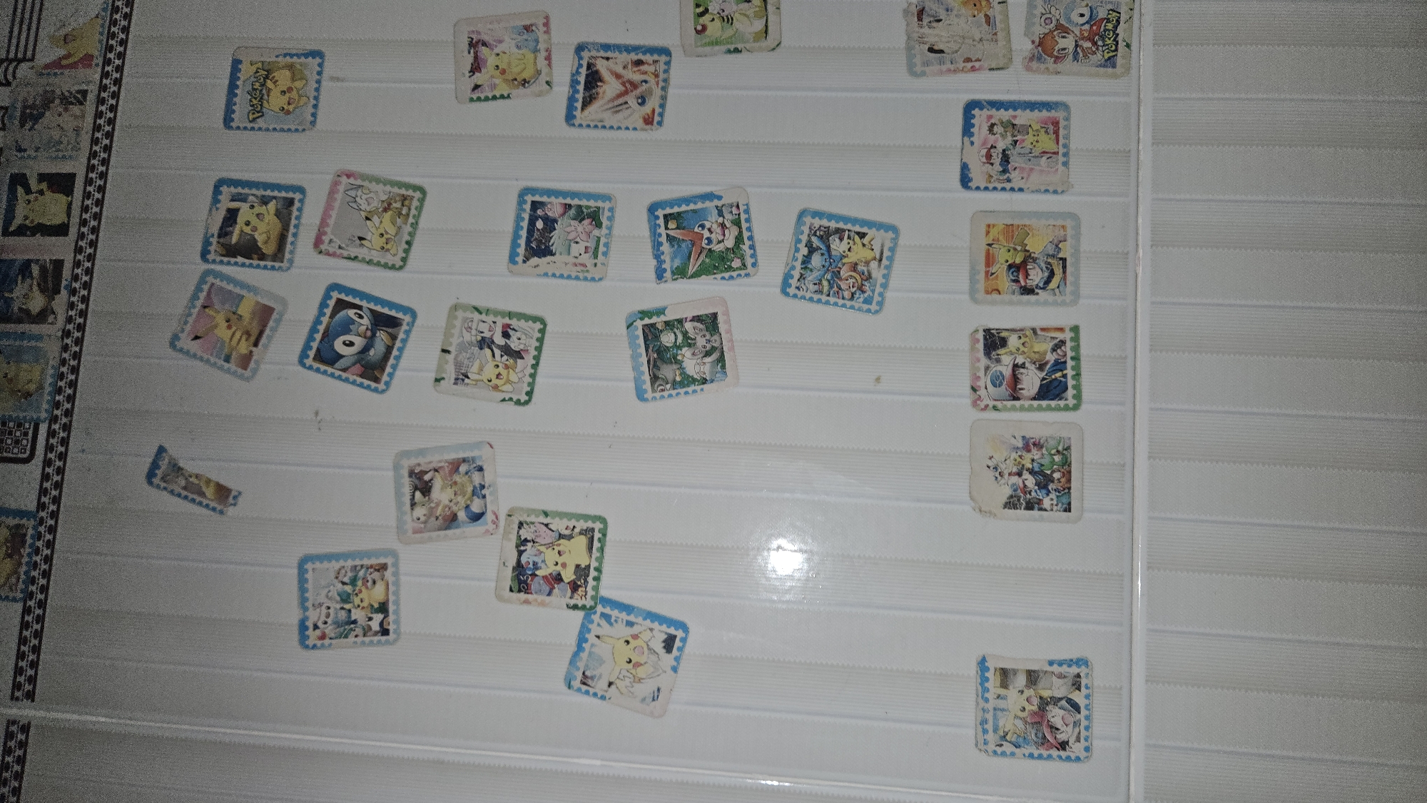 A collection of small, square stickers featuring Pokemon characters are scattered across a white, horizontally-ridged surface. The stickers have various colored borders (blue, pink, green) and depict different Pokemon characters and scenes. The white surface appears to be a wall or a board. The objects are generally arranged randomly, and they are present at different angles. The location is Cam Đức, Vietnam.