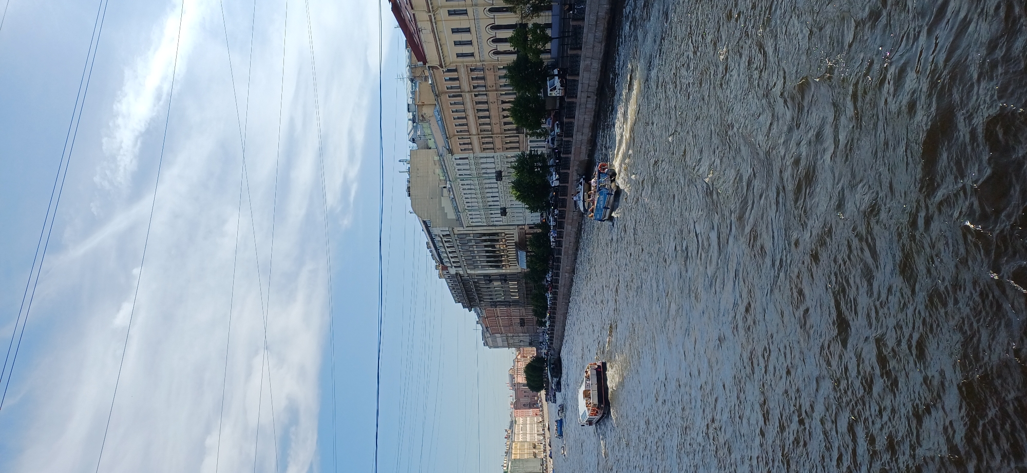 This is a photograph of a river in Saint Petersburg, Russia, likely during the daytime under a partly cloudy sky. The river is wide and choppy, with several boats on its surface. On the left side of the river, a large, rectangular boat with a white top and a dark hull is moving away from the viewer. On the right side, a smaller boat with a blue hull and a white top, carrying passengers, is moving towards the viewer. The riverbanks are lined with ornate buildings, characteristic of Saint Petersburg's architecture, with multiple stories and detailed facades. Trees are visible along the riverbanks between the buildings and the water. Overhead, multiple parallel wires are stretched across the frame, suggesting the presence of power lines or telecommunication cables. The overall scene depicts a busy urban waterway with architectural grandeur.