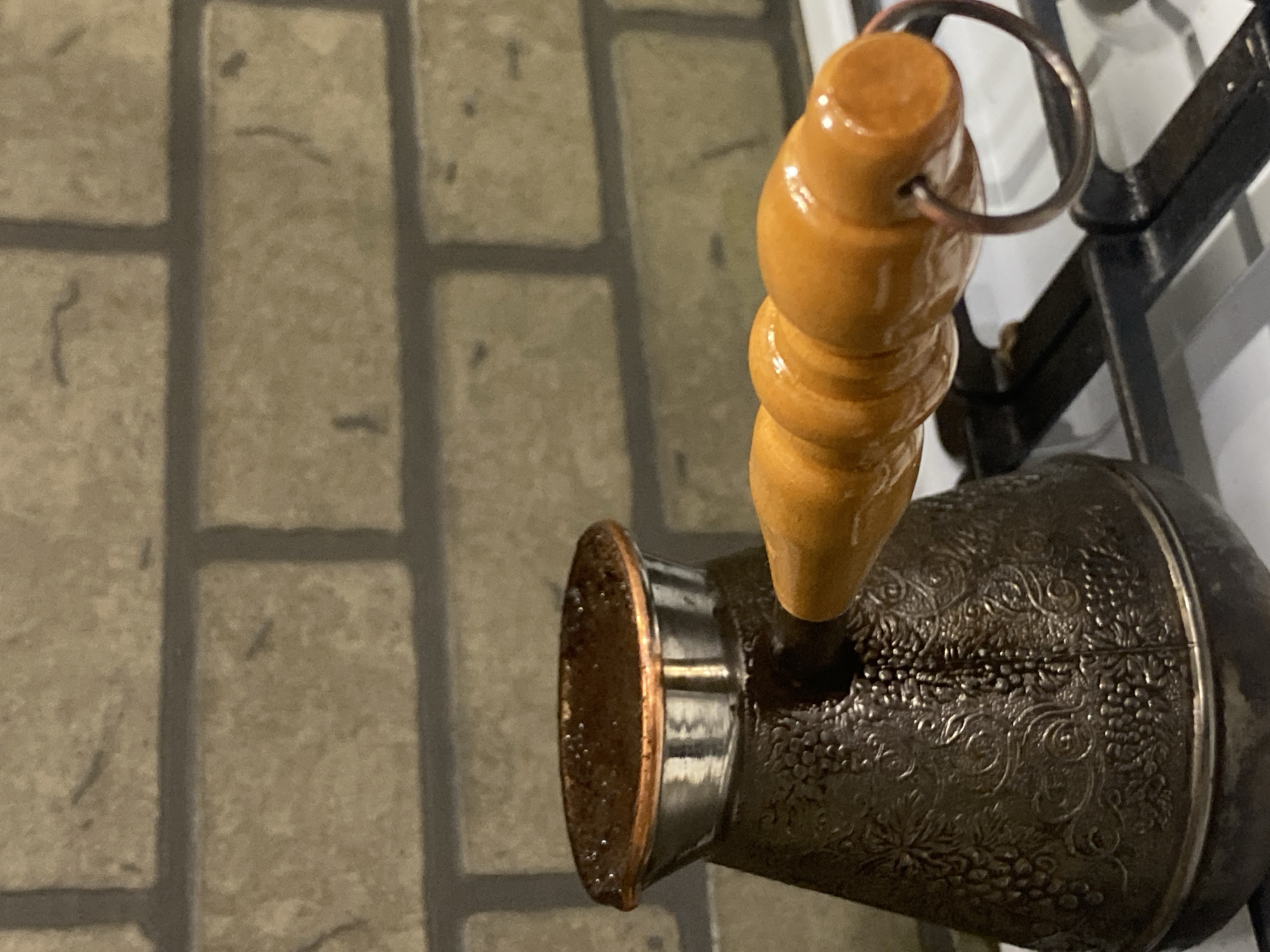 A close-up image shows a Turkish coffee pot (cezve) with a turned wooden handle resting on a stove grate. The pot has an ornate, possibly silver, exterior with floral designs. The handle is light brown. The background shows a tiled surface. The location is not discernible from the image.