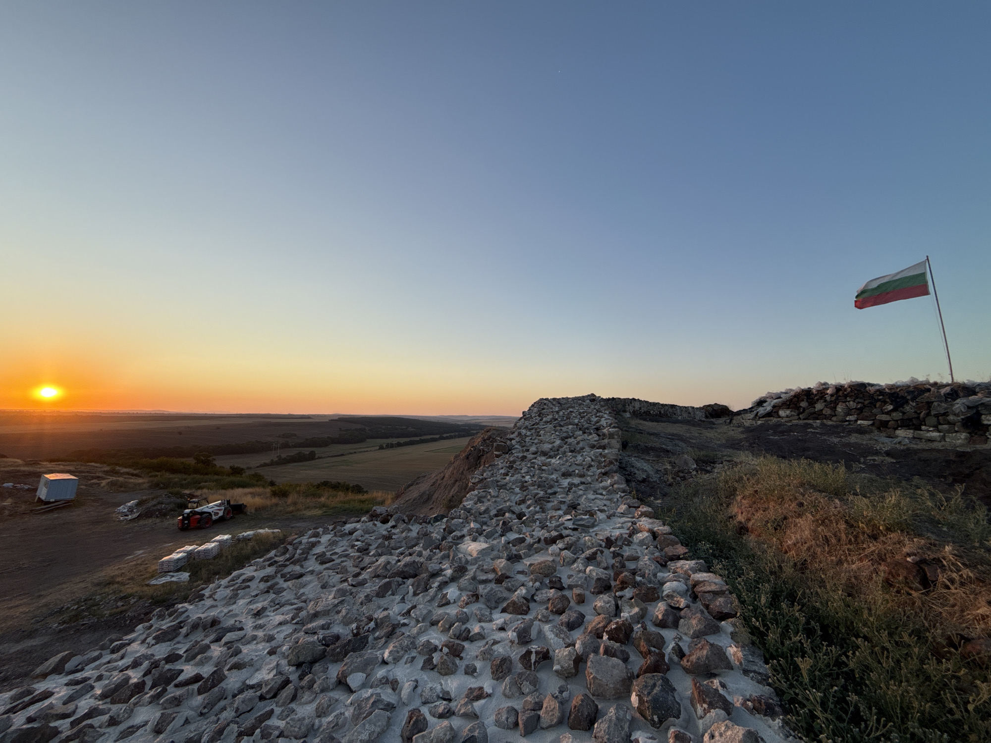The image captures a panoramic view from a hilltop fortification, likely the Rusokastro Fortress, in Rusokastro, Bulgaria, at sunset. The foreground is dominated by a rough, stone wall construction, with a path or road made of rocks and sand. To the left, a white portable structure, a skid steer loader, and stacks of building materials are visible on a dirt embankment, suggesting ongoing construction or restoration work. A wide, green landscape stretches out in the middle ground, featuring fields and trees, and beyond that, a distant horizon under a clear, gradient sky transitioning from warm orange at the horizon to a pale blue overhead. A Bulgarian flag is prominently displayed on a flagpole on the right side of the frame, waving in the breeze. The low angle of the setting sun casts a warm glow across the scene, highlighting the textures of the stone and the details of the surrounding landscape. The overall impression is one of historical significance juxtaposed with contemporary activity.