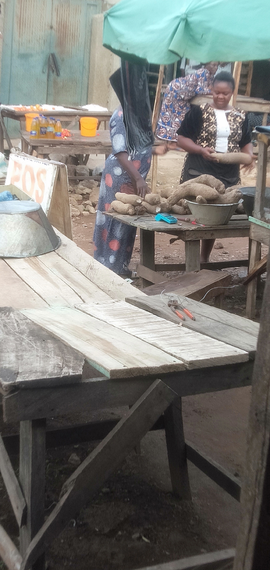 This image captures a vibrant outdoor market scene in Sabon Tasha, Nigeria, focusing on the sale and preparation of yams. Two women are prominent in the midground, positioned around rustic wooden tables filled with produce.
The woman on the right, wearing a patterned black top, holds a large yam, appearing to inspect or prepare it. Her expression is neutral to slightly pleasant. To her left, another woman, dressed in a blue and orange polka-dotted gown, stands near a heap of yams on a wooden table. Her head is partially obscured by a black bag or fabric hanging from a large green market umbrella that provides shade over the area. A large metal basin holds more yams on the table, and a small, bright blue electronic device, likely a payment terminal, is visible nearby.
In the background, a third woman in a patterned dress is partially visible, bending over. Another wooden table holds several plastic bottles of yellow liquid and two yellow plastic buckets. The backdrop consists of weathered corrugated metal walls, suggesting a permanent market structure or stalls. The ground is packed earth.
In the foreground, a bare wooden table displays a pair of orange-handled pliers and some wires. An inverted metal basin also rests on a lower foreground table. A white sign with the partial orange text "FOS" is leaning against a table on the left. The lighting indicates it is daytime, with the umbrella providing cover from direct sun. The scene encapsulates typical daily market activities focused on fresh produce.
