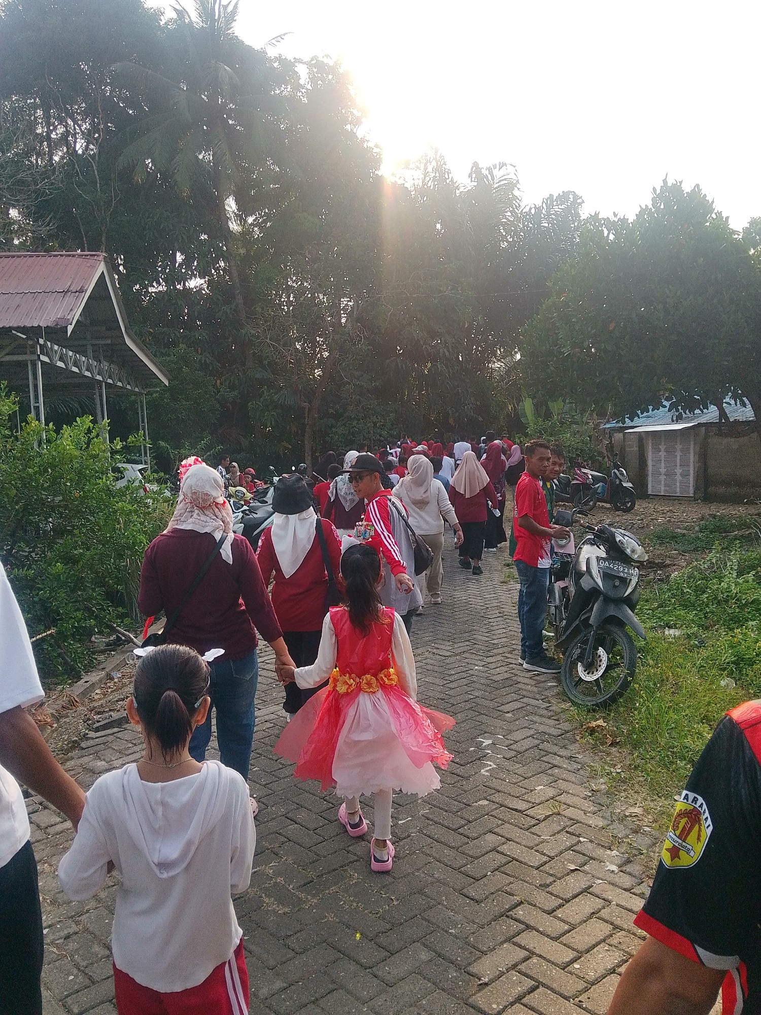 This is a medium shot, taken from a slightly elevated angle, of a group of people walking on a paved brick pathway. The setting appears to be a semi-rural area in Barabai, Indonesia, with lush green trees and vegetation lining the pathway. In the background, a simple building with a red tiled roof and a structure with white louvers are visible. Several motorcycles are parked along the side of the pathway.
The group consists of individuals of various ages. In the foreground, a young girl wearing a bright pink dress with yellow floral accents is being led by the hand by an adult. The adult, facing away from the camera, wears a maroon hijab and a matching maroon top. To the left of the young girl, another child in a white hoodie and red shorts is also walking, holding hands with someone whose arm is visible on the far left of the frame. Other people are walking in the same direction, some wearing headscarves and casual clothing. The sunlight is bright and appears to be either early morning or late afternoon, creating a lens flare effect in the upper left portion of the image. A man in a red t-shirt and blue jeans is standing by a motorcycle on the right side of the pathway, looking towards the camera and smiling. Another person partially visible on the far right wears a black shirt with a circular emblem on the sleeve. The overall atmosphere suggests a casual gathering or procession.
