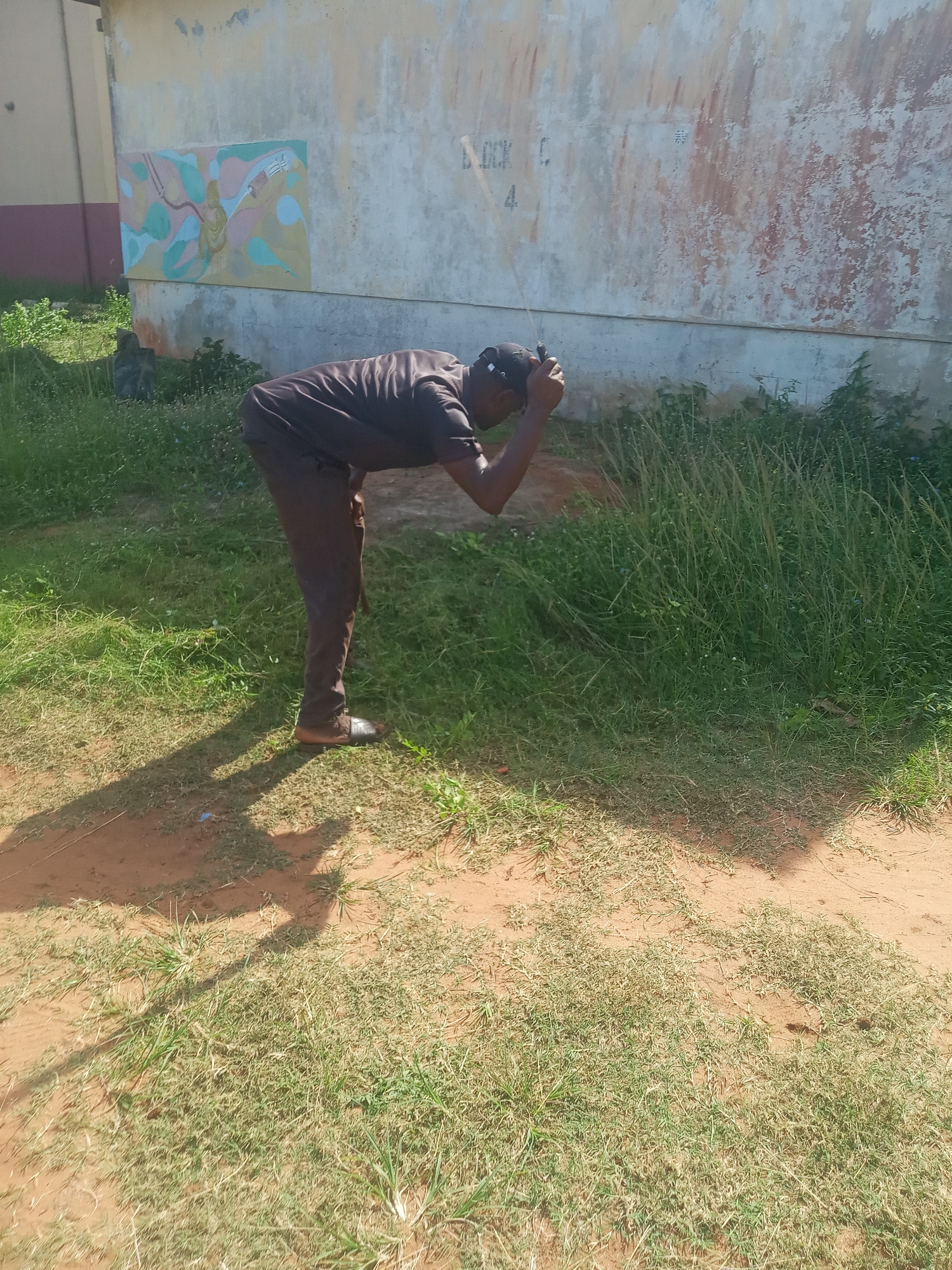 An outdoor, eye-level shot captures a man bent over in a sunny, grassy area next to a weathered building wall in Nsugbe, Nigeria. The man, dressed in a dark short-sleeved top, dark trousers, and dark slip-on shoes, has his head down and hands near his head, appearing to be focused on something in the tall green grass. His body is angled towards the right side of the frame.
The ground consists of reddish-brown dirt interspersed with patches of green grass, ranging from short to overgrown and wild. Strong, distinct shadows cast by the man and unseen objects indicate bright daylight, suggesting it is midday or early afternoon with clear weather.
In the background, a light-colored, possibly concrete, wall of a building spans across the frame. To the left of the man, a rectangular, abstract mural with soft, pastel colors (blue, pink, yellow, green) is affixed to or painted on the wall. Further left, a portion of an adjacent building with reddish-brown and darker-colored sections is visible. On the weathered wall to the left of the man, faint text reads "BLOCK C" and the number "4". The wall shows signs of age, with peeling paint and discoloration.