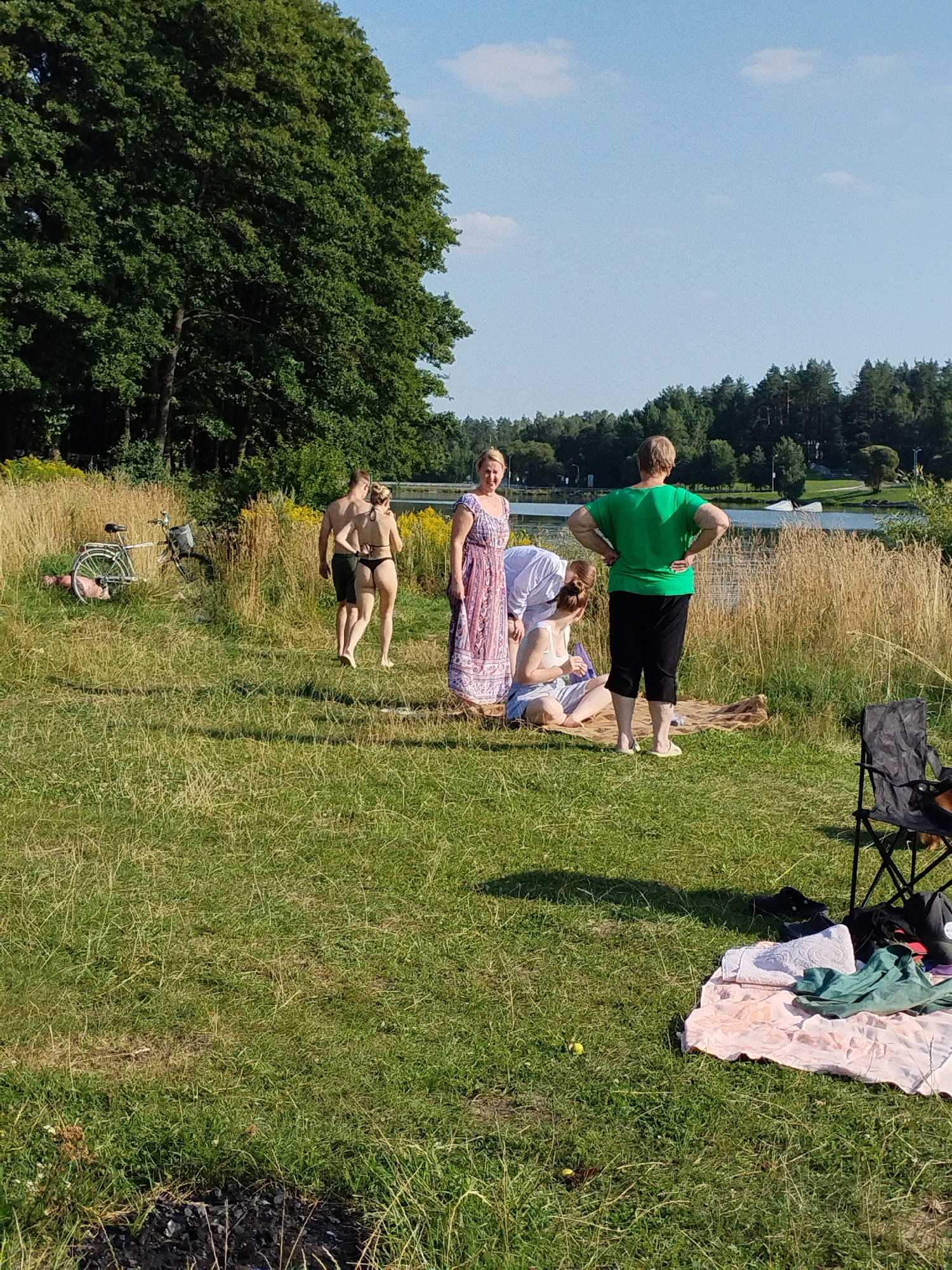 The image depicts a sunny day at a lakeside, likely in or near Навадворскі сельскі Савет, Belarus, given the presence of lush greenery and a body of water. Several individuals are present, engaged in various activities. On the left, a man and a woman in a bikini are walking away from the camera, their backs to us. Nearby, a bicycle is leaning against the tall dry grass, and a pink object, possibly a towel or blanket, is on the ground next to it. Towards the center-right, another group of people are gathered on the grass. One woman in a long, patterned dress stands looking towards the lake, while another woman in a green t-shirt and black shorts stands with her hands on her hips, also facing away from the camera. Two other individuals are partially visible, one seated on a blanket and another bending over. In the foreground, a black camping chair is set up, with various items of clothing and bags on a pink blanket beside it. The environment is a grassy field with tall, dry vegetation bordering a lake. The background shows a dense line of trees and a clear blue sky with a few scattered clouds. The overall impression is a casual, outdoor gathering near a lake on a pleasant day.