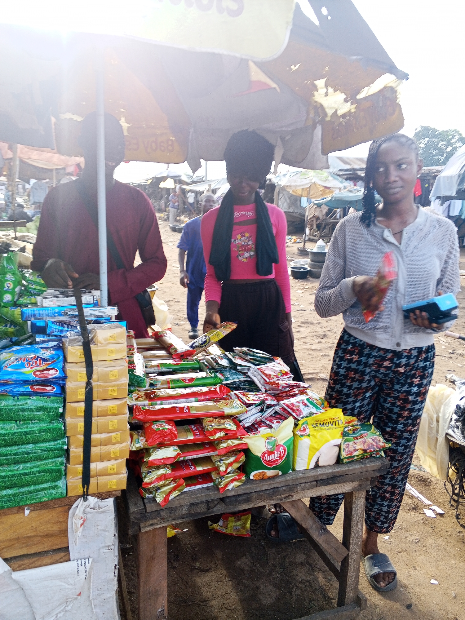 In Lokoja, Nigeria, a stall displays various goods under a large, partially damaged yellow and white umbrella. A man in a maroon shirt stands behind the stall, attending to the merchandise. To his right, a woman in a grey, mesh-like cardigan and patterned pants holds a red packet in her right hand and a blue electronic device in her left, appearing to conduct a transaction. Behind her, another woman in a pink top and dark skirt stands with a black scarf around her neck. The stall is laden with packaged food items, including what appear to be pasta, seasonings, and other dry goods, arranged on wooden shelves and stacked high. The ground is dirt, and other stalls and market structures are visible in the background under an overcast sky.