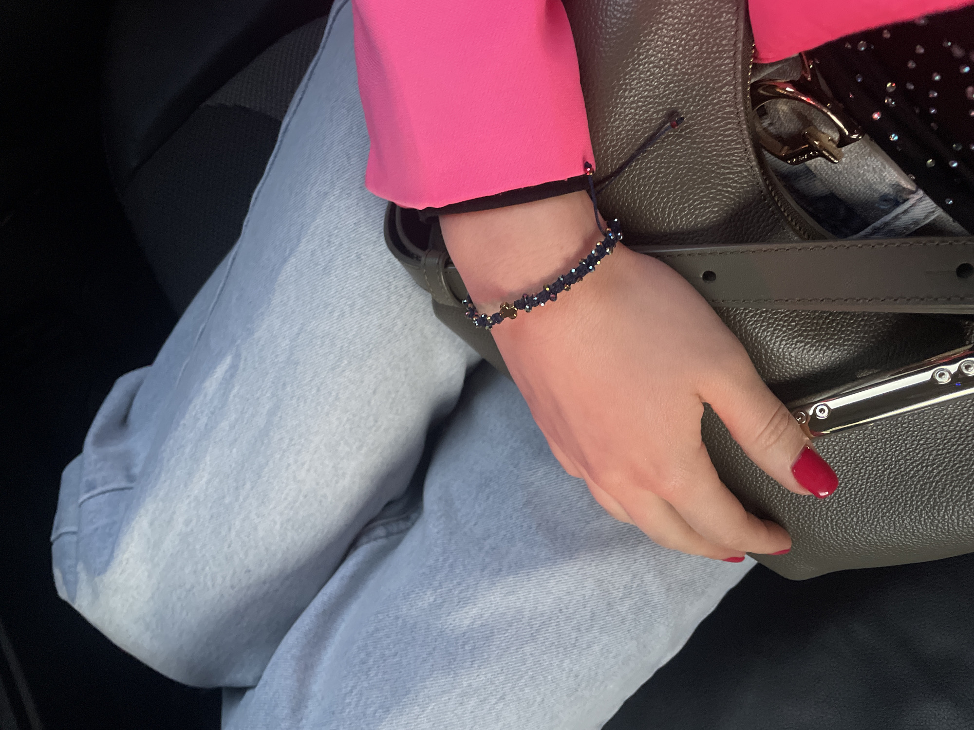 A woman's left hand rests atop a green leather handbag. She is wearing light blue jeans and a hot pink blazer with a black trim at the cuff. A dark blue beaded bracelet adorns her wrist. The setting appears to be inside a vehicle, with a dark car seat visible in the background. The location is not possible to identify.