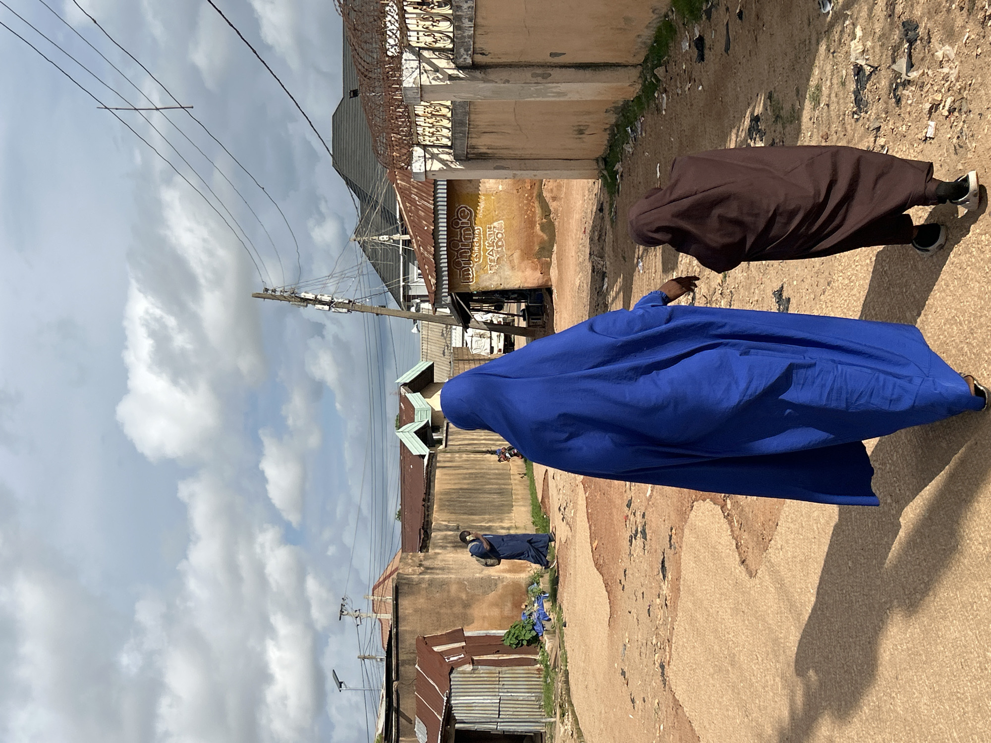 In the city of Jos, Nigeria, a street scene unfolds under a partly cloudy sky. Two individuals, one an adult and the other a child, are walking away from the camera, their figures draped in dark brown and vibrant blue coverings, respectively. The adult, in blue, appears to be leading, with the child in brown following closely behind. The ground beneath them is a dusty, dirt road, marked by tire tracks and scattered debris.
To the left of the road, a corrugated metal structure with a rusted roof stands beside a more substantial building with a tan-colored wall. Green foliage sprouts near the metal structure. Further down the road, another individual, also in dark blue attire and carrying a backpack, walks towards the camera, appearing to be engaged in a phone conversation.
The background reveals a cityscape characterized by various rooflines, including corrugated metal and darker tiled roofs, suggesting a mix of residential and commercial structures. Utility poles with an array of wires crisscross the sky, a common sight in urban environments. A sign with text, partially obscured and in an unknown language, is visible on one of the buildings. The overall impression is one of a typical day on a bustling, yet somewhat weathered, street in Jos.