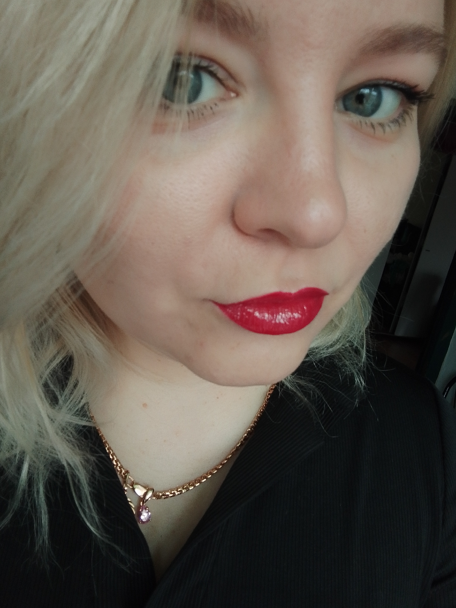 A close-up, chest-up image depicts an individual, likely female, situated in the city of Izhevsk, Russia. The subject has blonde, wavy hair framing a fair-skinned face. Her left eye (viewer's left) is partially obscured by hair, while her right eye (viewer's right) is fully visible, featuring a light blue-green iris, dark pupil, and visible eyelashes with mascara. Her lips are full, coated in a glossy, vibrant red lipstick, and positioned in a slight pout. She wears a black garment, appearing to be a ribbed fabric top or sweater. Around her neck is a gold-toned chain necklace with a small pendant containing a pink, tear-shaped gem. The background is dark and out of focus, indicating an indoor setting, with vague vertical lines visible to the right. The individual gazes directly towards the camera.