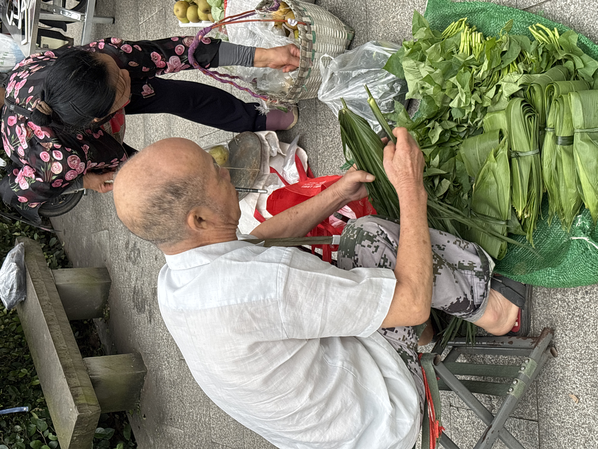 In Jiangjin, China, an elderly bald man wearing a white linen shirt and camouflage pants sits on a folding stool, preparing bundles of green leaves. He is in the process of wrapping more leaves around a stalk. To his right, a woman wearing a black top with a floral pattern and dark pants is bent over, placing items into a woven basket. The basket contains what appear to be pears and other produce. A pile of green vegetables, including bundled leafy greens and other items wrapped in leaves, sits on a green mat in the foreground. The scene takes place outdoors on a paved surface, with a stone bench and a leafy hedge visible in the background.