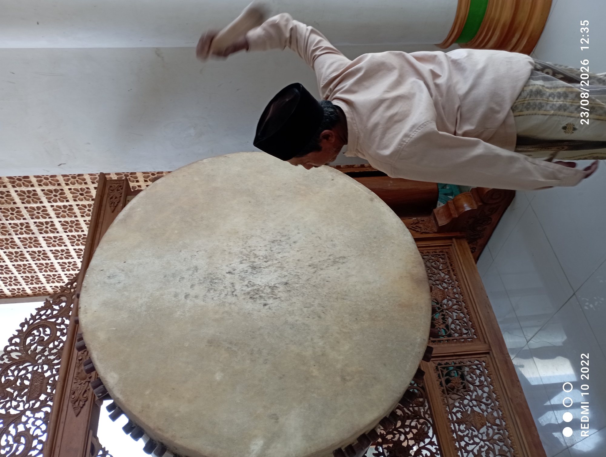 In Dukuhseti, Indonesia, an elderly man wearing a black peci hat and a light-colored long-sleeved shirt is captured in motion, striking a large drum. The drum, with a pale, textured surface, is supported by an intricately carved wooden frame, suggesting cultural or religious significance. The man's right arm is raised, mid-swing, implying he is actively playing the instrument. The background features a white wall and a patterned decorative screen, hinting at an indoor or sheltered environment, possibly a place of worship or a cultural center. The floor appears to be tiled. A date and time stamp in the lower right corner indicates the image was taken on 23/08/2026 at 12:35.