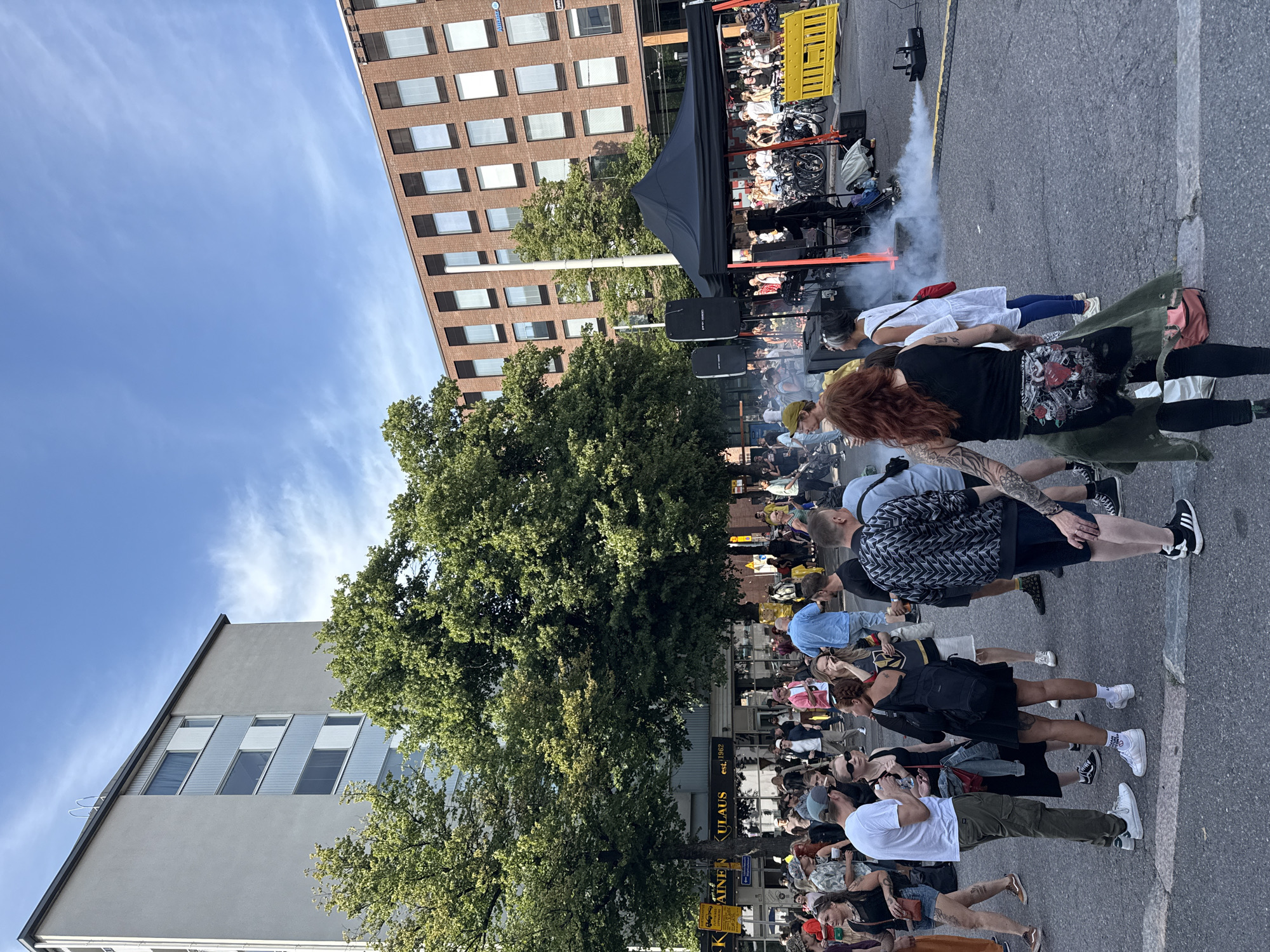The image depicts an outdoor event in Helsinki, Finland, likely in a public square or street. Several people are gathered, with some appearing to be participants and others observing. In the foreground, a group of individuals is seen milling about, engaged in conversations and casual interactions. There are trees and buildings visible in the background, suggesting an urban setting. One building is a multi-story brick structure, while another appears to be a more modern, lighter-colored building. A black tent is set up, possibly for a performance or vendor. Smoke is emanating from a machine near the tent, creating a hazy effect. The ground is paved, indicating a street or plaza. The overall atmosphere appears to be that of a lively public gathering.