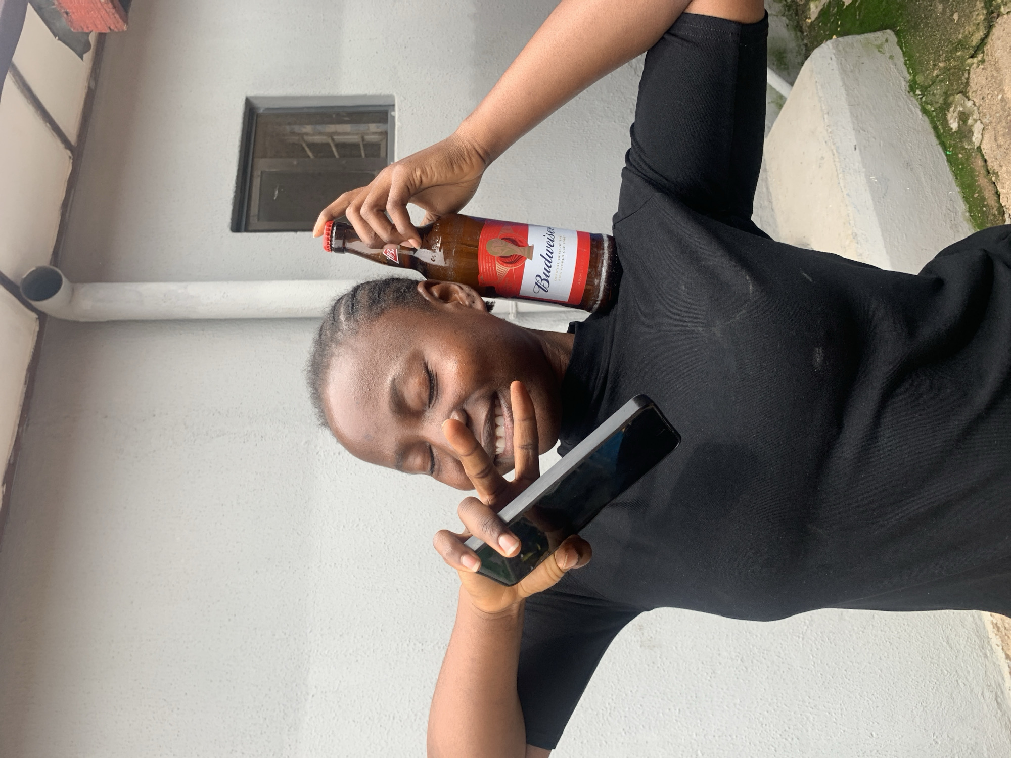 A young Black woman with braided hair is holding a bottle of Budweiser beer on top of her head while smiling and making a peace sign with her fingers. She is also holding a black smartphone in her other hand, positioned in front of her face. She is wearing a black t-shirt. The background consists of a light gray wall with a small window and some green moss on a concrete structure. The scene appears to be outdoors, possibly in a courtyard or yard. The lighting suggests it could be daytime, and the presence of moss implies an environment with some moisture, potentially after rain or in a damp area. The woman's expression is joyful and playful, indicating a lighthearted moment. The visible text on the beer bottle reads "Budweiser" and "OFFICIAL BEER OF THE FIFA WORLD CUP". The overall impression is one of fun and relaxation.