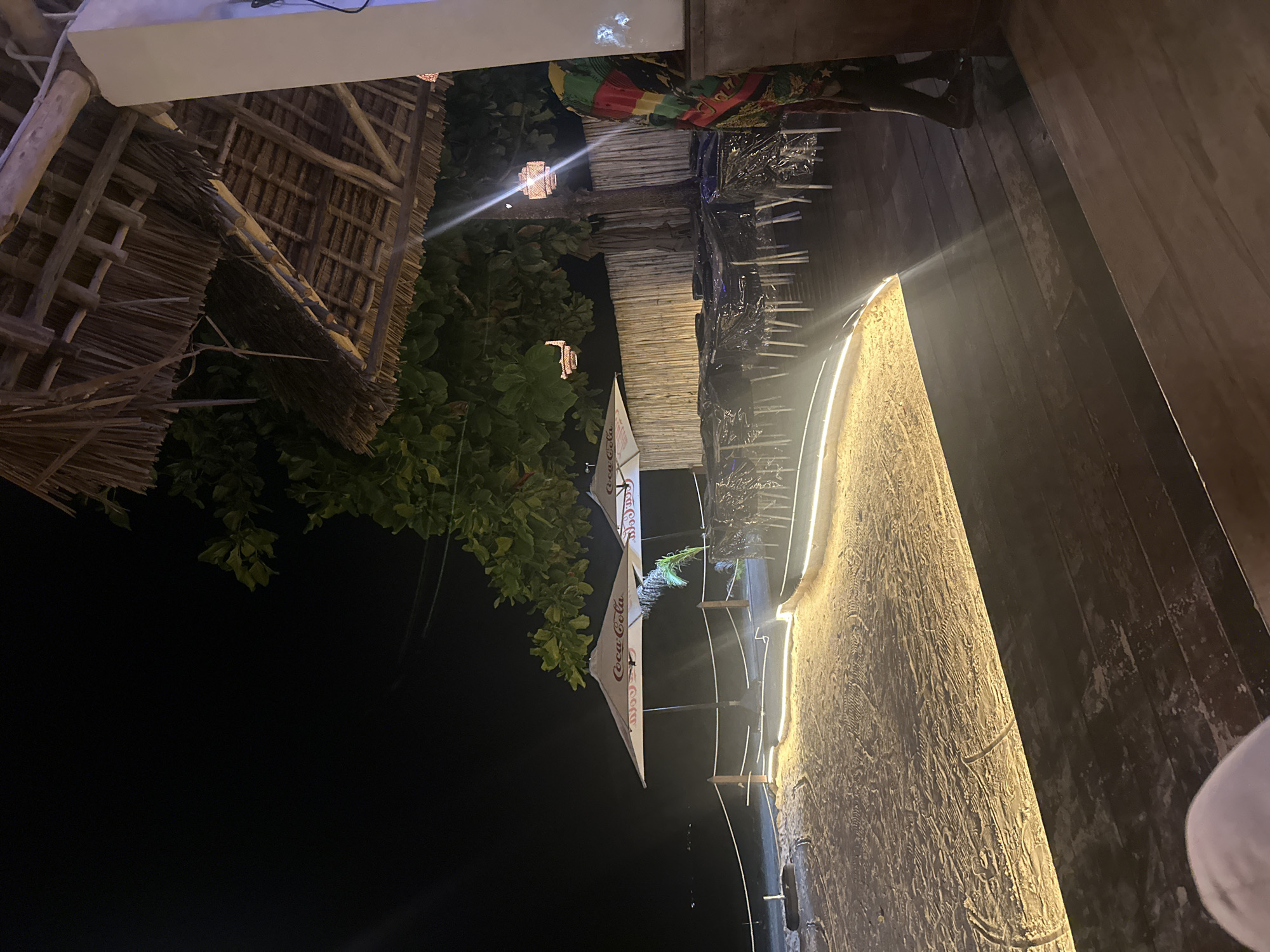 The image appears to be taken in a restaurant or bar area in an unidentified city. The scene seems to be at night, with a mix of natural and artificial lighting. The main elements include a thatched roof structure, possibly part of a bar or outdoor seating area, adorned with green foliage. There are Coca-Cola branded umbrellas providing shade. The floor is wooden and has built-in lights along the edge. There are chairs and tables, indicating a dining or lounging area. The surroundings are dark. The orientation of the image has the horizon running vertically.