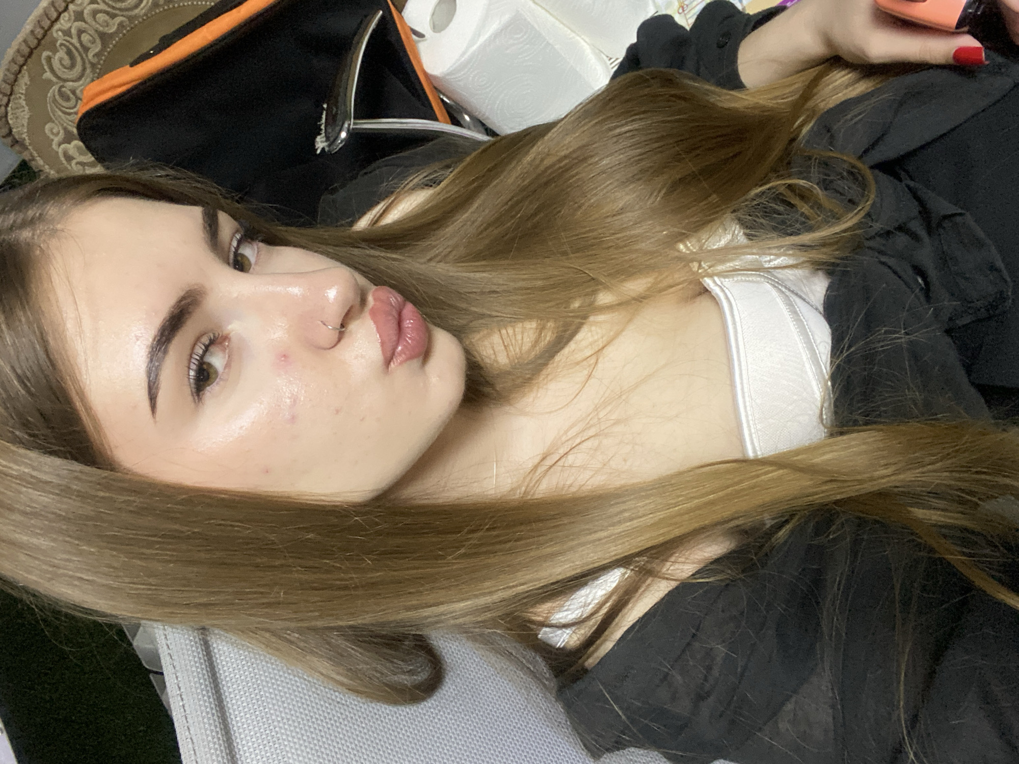 The image is a close-up, waist-up, selfie-style photograph of a young Caucasian woman with long, light brown hair. She is wearing a white top with a textured pattern and a dark, open-front garment. She has a nose ring and is puckering her lips. Her skin appears to have some minor blemishes. In the background, there is a grey upholstered surface on the left and a dark bag with an orange trim on the right, along with rolls of white paper towels. The overall environment suggests an indoor setting, possibly a room or a vehicle, but there are no specific landmarks to confirm a location within Dyakovo, Russia.