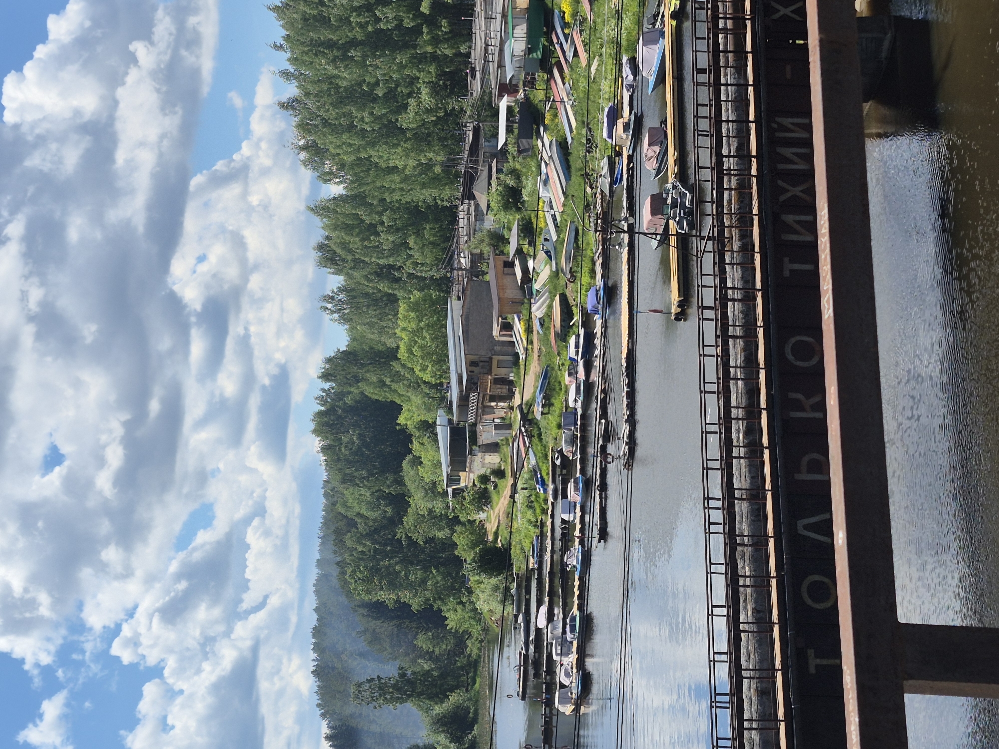 The image presents a scene in Chaikovsky, Russia. On the right, a body of water reflects the cloudy sky. Along the bank, there are numerous boats docked near a tree-lined area with a few buildings. A structure with Cyrillic text, "ТОЛЬКО ТИХИЙ X" is visible on the lower-right side of the image.