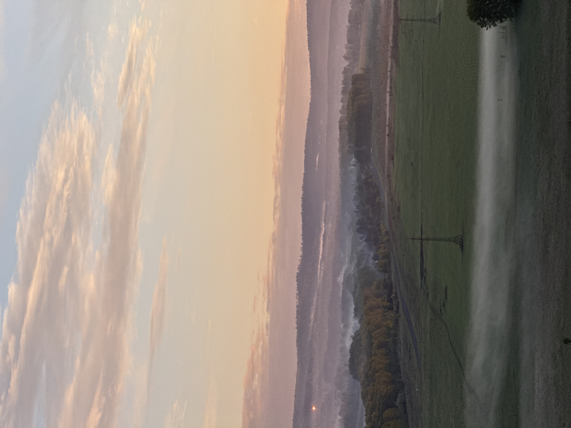 The image presents a landscape view during either sunrise or sunset. On the left, the sky transitions from light blue to orange with scattered clouds. To the right, a series of hills or small mountains are visible, enveloped in a hazy fog. At the base of the hills, a strip of trees is visible. Adjacent to the hills, there's a flat green field with power lines cutting across it. Fog stretches over the field, obscuring the details of the ground. There is no information in the image to indicate the location is Shamoni, Russia.