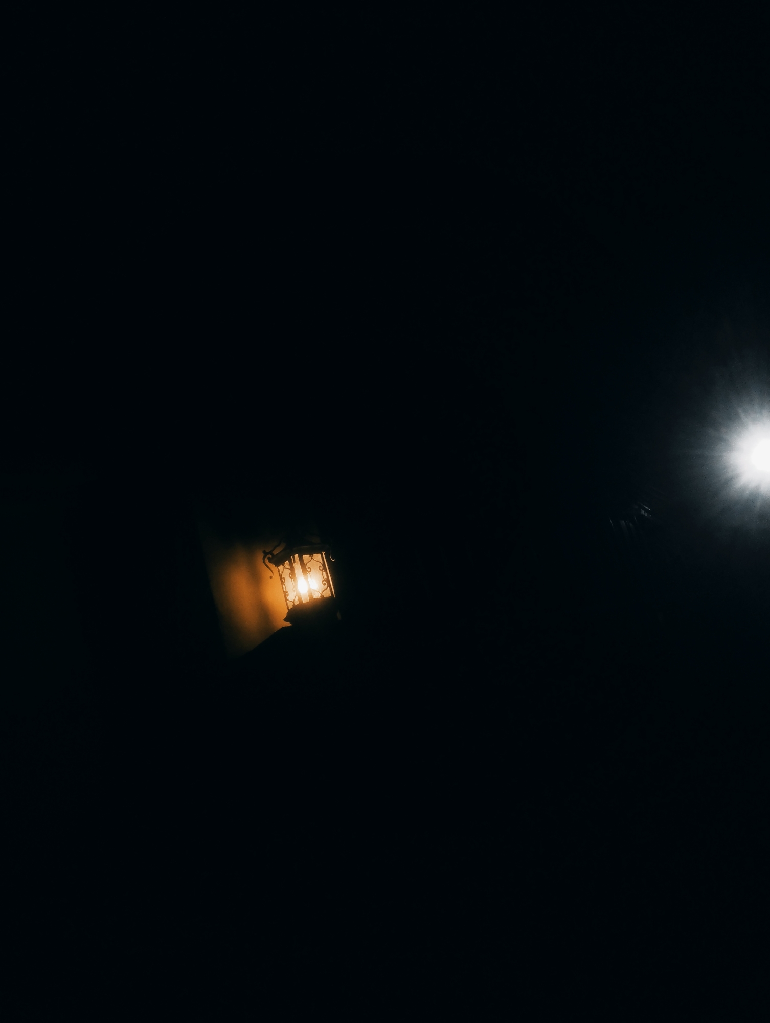 A dark, atmospheric image features a vintage-style lantern emitting a warm, orange glow from its illuminated interior. The lantern is positioned to the left of the frame, casting a soft light on a dark, textured surface behind it. To the right, a bright, starburst-shaped light source, possibly a distant streetlight or a lens flare, pierces the darkness, creating a high-contrast scene. The overall impression is one of nighttime in an urban setting, with the interplay of light and shadow dominating the composition. The specific location in Jos, Nigeria, is not discernible from the visual information presented.