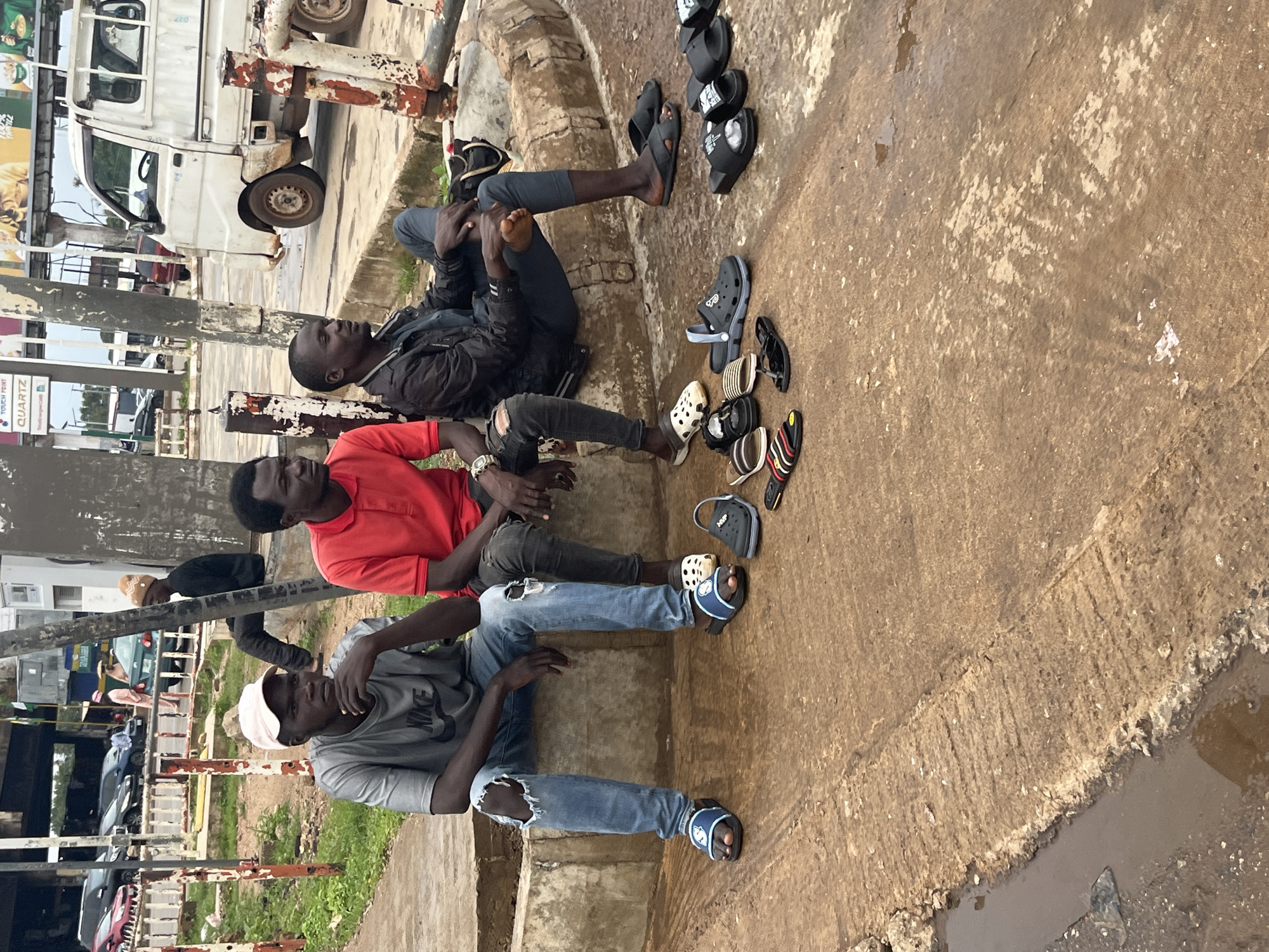 Three young men are sitting on a concrete ledge in what appears to be an outdoor urban environment, possibly in Jos, Nigeria. The man on the left is wearing a grey t-shirt and ripped blue jeans, with a pink baseball cap. He is resting his chin on his hand. The middle man wears a red t-shirt and dark pants. The man on the right wears a dark jacket over a t-shirt and dark pants. He is looking to his left. A variety of footwear, including sandals and slides, are scattered on the ground in front of them. In the background, a white truck and other vehicles are visible, along with some buildings and greenery. The ground in front of the men is wet, with puddles of water on the reddish-brown concrete. There are also some metal posts and a concrete barrier visible.