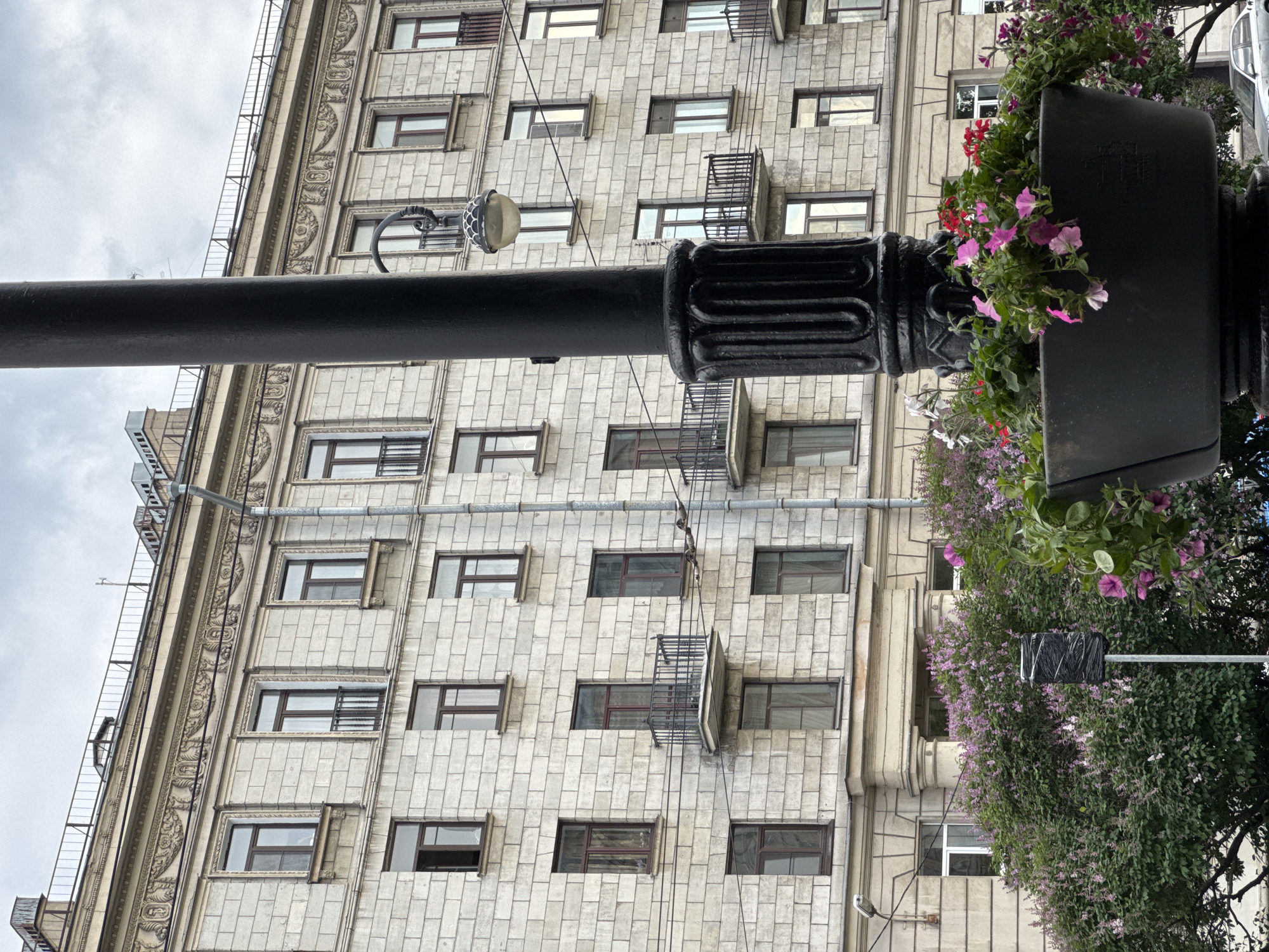 The image displays a daytime urban scene, dominated by a large, ornate black lamppost extending diagonally from the lower left to the upper right. A decorative planter box is affixed to the lamppost, spilling over with vibrant pink, red, and white petunia-like flowers and green foliage.
In the background, a grand, multi-story building rises, its facade constructed from light-colored stone, possibly granite or limestone, laid in a distinct block pattern. The building features numerous rectangular windows, some with dark frames, and several are adorned with small, decorative black iron balconies or window guards. Intricate architectural details, including carved motifs and moldings, are visible along vertical sections of the building's exterior. A smaller, ribbed metal lamp is also attached to the main lamppost.
The sky above the building is overcast, indicating a cloudy day. Green trees and bushes are visible behind the lamppost and to the right, adding natural elements to the streetscape. No specific activity or event is taking place, presenting a static view of the city. The architectural style, with its grandeur and decorative elements, is consistent with historic buildings found in Saint Petersburg, Russia. No clearly legible text is visible on any surface within the frame.