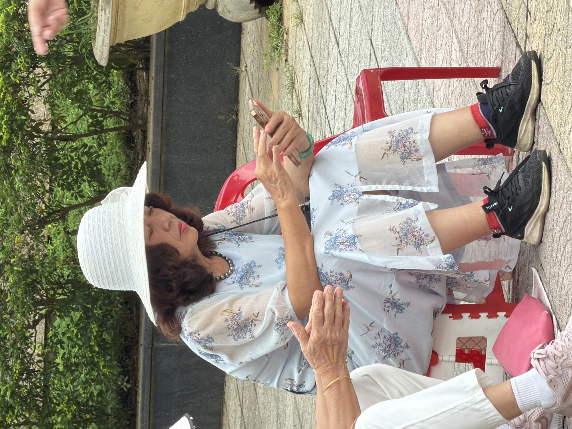 An elderly Asian woman wearing a white sun hat and a light blue floral dress is seated on a red plastic chair outdoors. She is holding a smartphone in her hands and appears to be looking at the screen. Next to her, another person's hands are visible, with one wearing a gold bracelet and the other showing signs of age. In the foreground, a pink cushion and a white collapsible stool are on the ground. The woman is wearing black sneakers with red socks. The background consists of green foliage and a large decorative planter. The scene appears to be in a public outdoor space in Thuận Hóa Ward, Vietnam, with the ground paved with square tiles.
