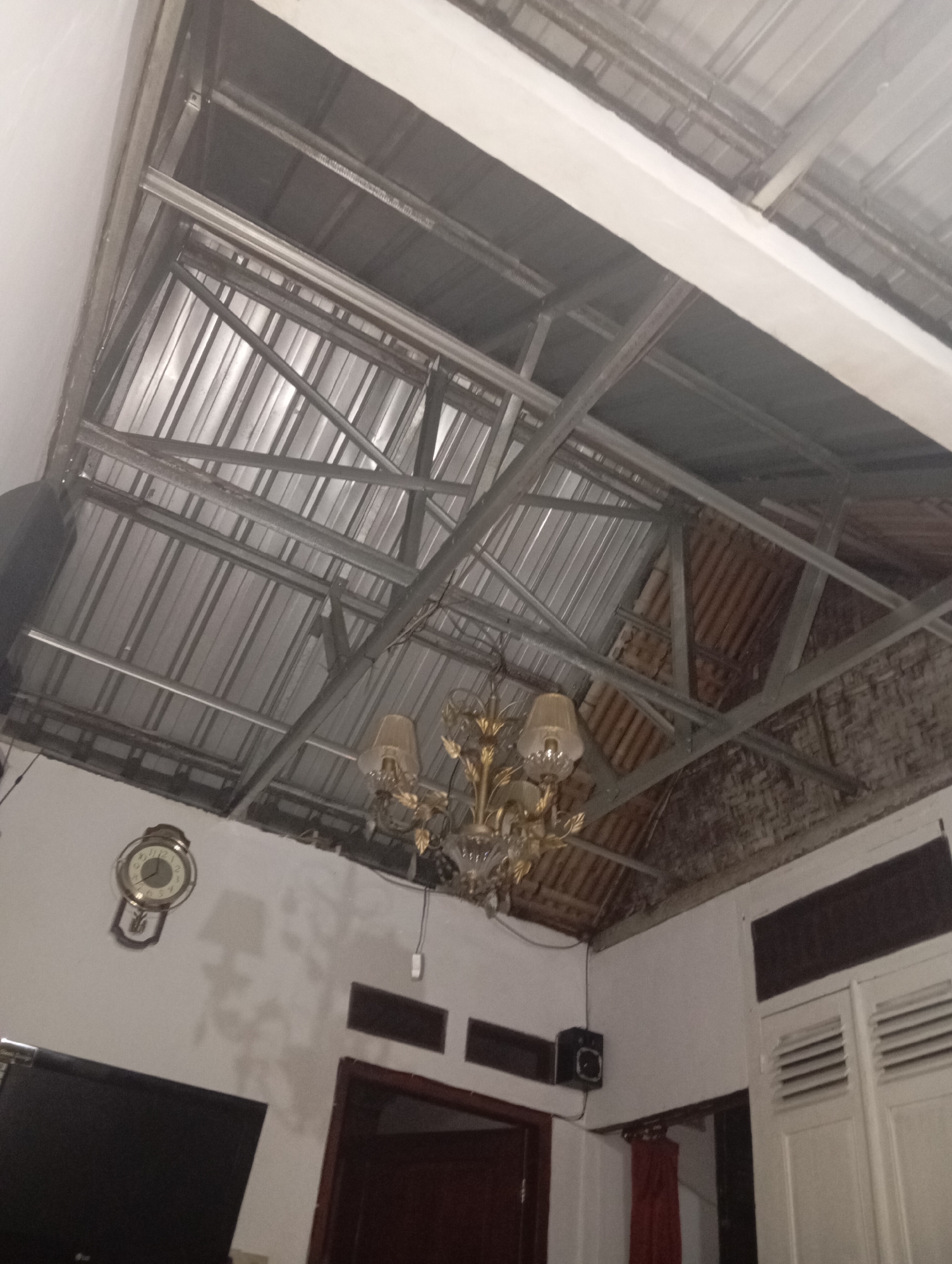 An interior shot captures a room with an unfinished ceiling. The ceiling structure is made of exposed metal beams and corrugated metal sheeting. A decorative chandelier with multiple glass shades hangs from the ceiling. On the wall to the left, a vintage analog clock with a wooden frame is mounted. To the right of the clock, a dark flat-screen television is visible. Further to the right, a white wooden louvered door or cabinet is present. The lighting in the room is dim, with shadows cast on the wall, particularly from the chandelier. The walls appear to be white. The overall impression is of a partially completed or rustic interior space.
