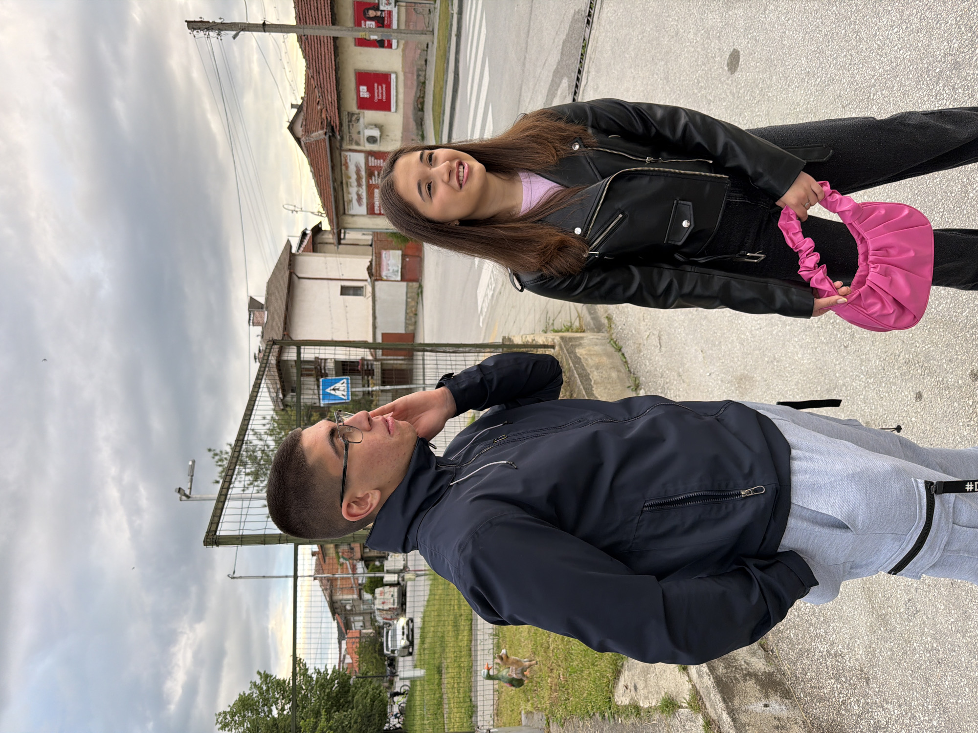 The image shows a young man and woman posing in front of a street scene in Panagyurishte, Bulgaria. The man is wearing glasses, a navy jacket, and grey sweatpants and appears to be looking upwards. The woman is wearing a black leather jacket over a lavender top with black jeans, and she is holding a bright pink handbag. A crosswalk and several buildings are visible in the background, including one with red signage. A metal fence and some greenery are visible near the man. The sky is overcast.