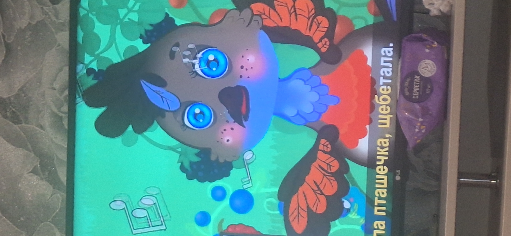 The image shows a cartoon bird character with large blue eyes and orange wings, against a green background with musical notes and berries. Below the cartoon, there is text in yellow, partially visible, reading "ла птичечка, щебетала." Below the screen, an LG logo is visible. Further down, a purple package of "СЕРБЕТКИ" (wipes) is placed on a shelf. The background appears to be a textured wall. There is no indication of the city of Bila Tserkva, Ukraine, in the image.