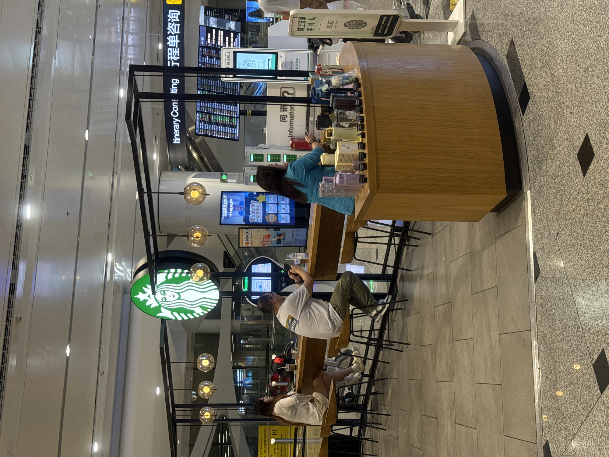 This image depicts a Starbucks coffee shop located in an airport terminal. The Starbucks logo, a green circular emblem with a siren, is prominently displayed above the counter. The shop features a wooden counter with several patrons seated on high stools. A woman in a light-colored outfit is seated on the left, and a man in a gray t-shirt and green pants is seated in the middle, both looking at their phones. A woman in a blue top is standing behind the counter, interacting with a customer. The counter is adorned with various merchandise, including reusable cups and water bottles. Behind the counter, a departure board with flight information is visible, indicating the airport setting. The floor is tiled with a neutral gray stone pattern. The overall environment appears to be a busy public space, typical of an airport concourse.