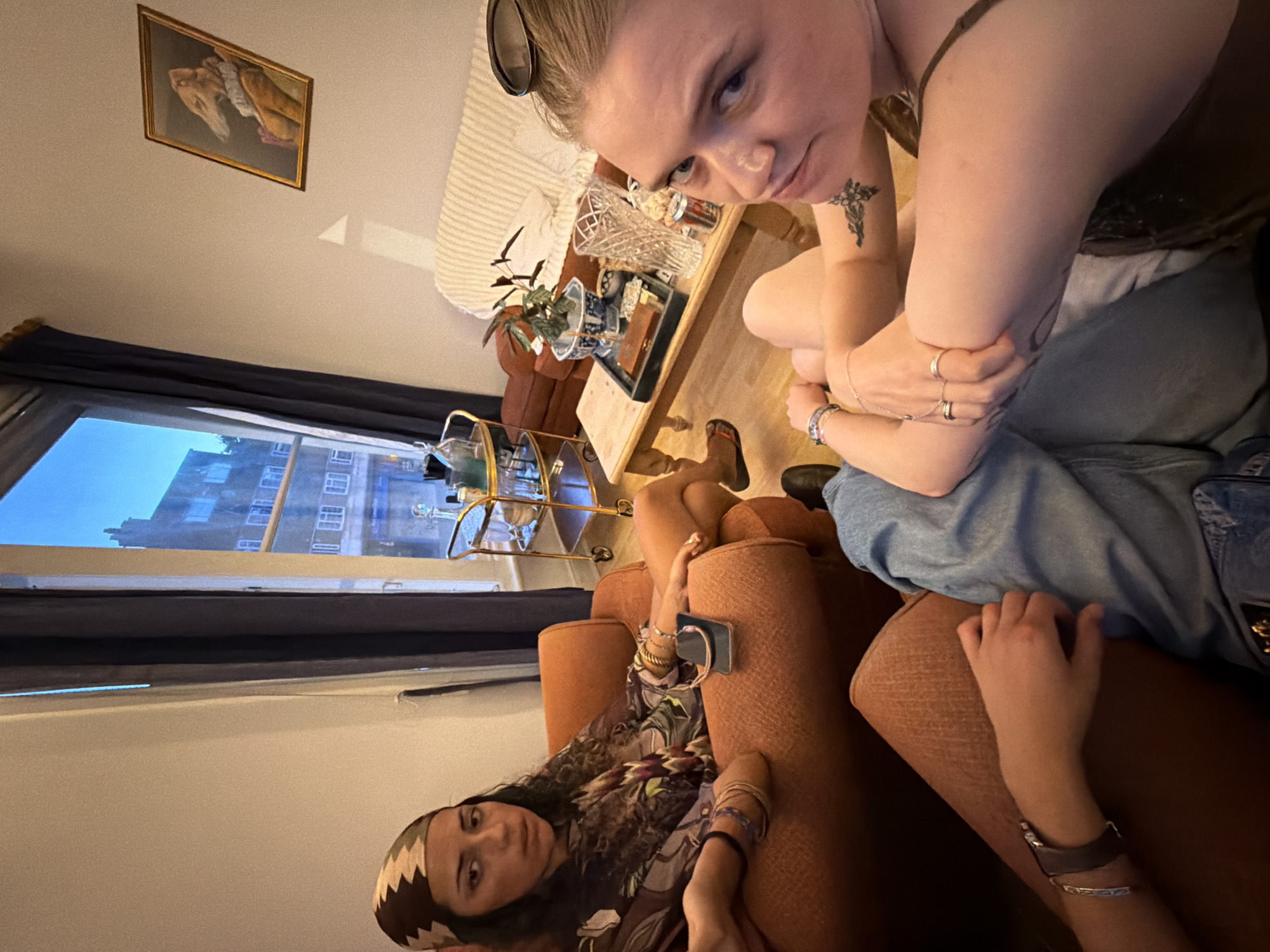 The image depicts a group of individuals indoors, likely in a residential setting, possibly a living room. The ambiance suggests a casual gathering.
One individual, wearing a patterned headscarf and a printed top, is seated in an orange armchair. They are looking towards the camera with a neutral expression. Their legs are crossed, and they are wearing bracelets on their left wrist. A phone is visible on the armrest of the chair.
Another individual, whose face is partially visible and angled towards the camera, is seated in the foreground. They appear to be wearing a dark top and have tattoos on their arms. Their arms are crossed, and they are wearing rings on their fingers.
A third person, only their legs are visible, is seated on the floor, wearing blue jeans.
The room contains a sofa with striped upholstery, a wooden coffee table with various items on it including a vase, a tray with what appears to be a box, and cans. A bar cart with bottles is positioned near a window. The window overlooks a building with multiple windows, suggesting an urban environment. A framed picture, possibly of an animal in attire, hangs on the wall. The lighting suggests it is evening or late afternoon, with natural light coming from the window and artificial light within the room.
The location is stated to be in the city of London, United Kingdom. Without further context or specific landmarks, it's difficult to pinpoint the exact neighborhood. However, the architectural style of the building visible through the window, and the general interior design, are consistent with many residential areas in London.