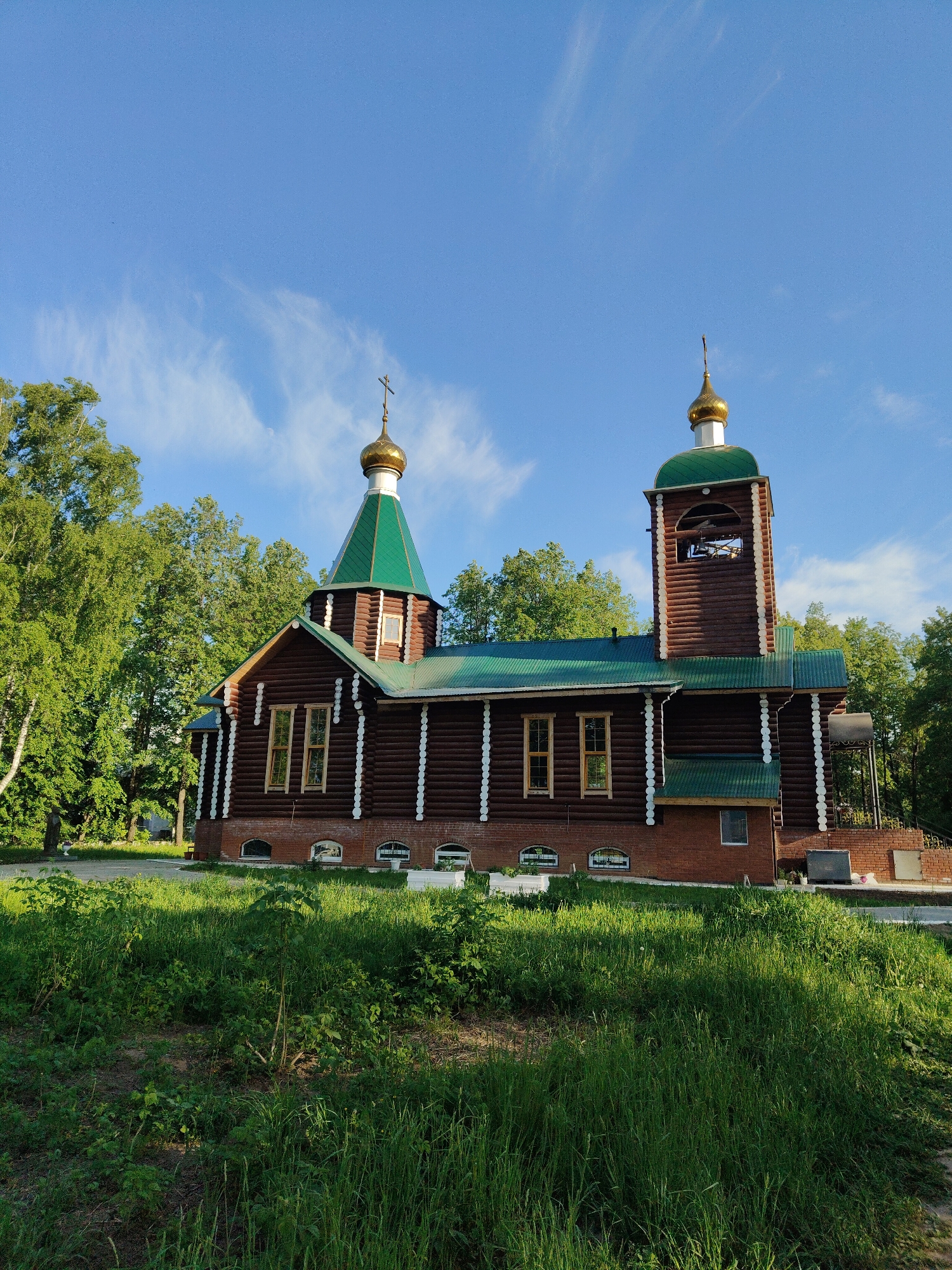 A wooden church with green roofs and golden domes stands in a grassy area under a blue sky with wispy clouds. The church is brown with white trim and brick base, and has multiple windows. Trees surround the church, and the foreground consists of green grass and vegetation. The location is Unknown, Unknown.