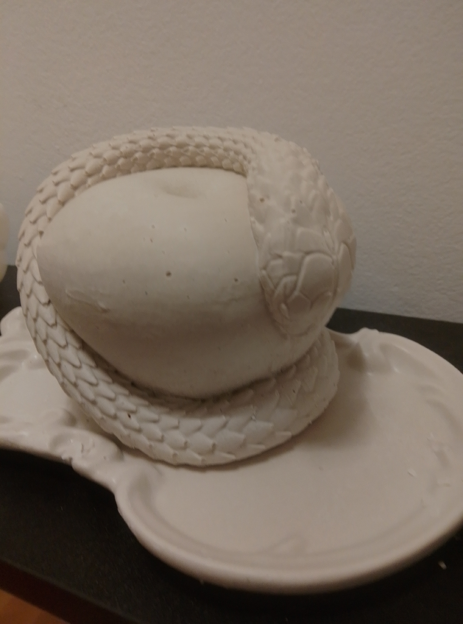 A white, unglazed ceramic sculpture of a coiled snake is displayed on a matching ceramic plate. The snake's body is textured to resemble scales, and its head rests on a smooth, egg-shaped form. The sculpture is positioned on a dark, flat surface, possibly a tabletop. The background consists of a plain, light-colored wall. There are no people or other animals visible in the image. The scene is indoors, with soft lighting. The location in the city of Wedel, Germany is not discernible from the image.