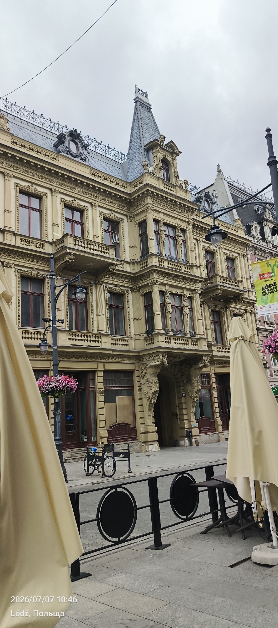 The image shows a section of a light yellow, ornate building with a dark grey, mansard roof and a pointed turret. The building features multiple stories with numerous windows, some of which have red frames. Decorative elements, including sculpted figures supporting balconies and intricate carvings, adorn the facade. In the foreground, there is a street with a paved sidewalk. A black metal railing with circular inserts separates the sidewalk from the street. To the left, a large, light yellow umbrella obscures part of the view. A lamppost with a hanging flower basket is visible. A bicycle is parked on the sidewalk. The bottom left corner of the image contains a timestamp and location: "2026/07/07 10:46 Łódź, Польша." A banner, partially visible on the right side of the building, displays text in green and white, with "mbank" and "ŁÓDŹ SUMMER FESTIVAL" being legible. The sky is overcast and grey.