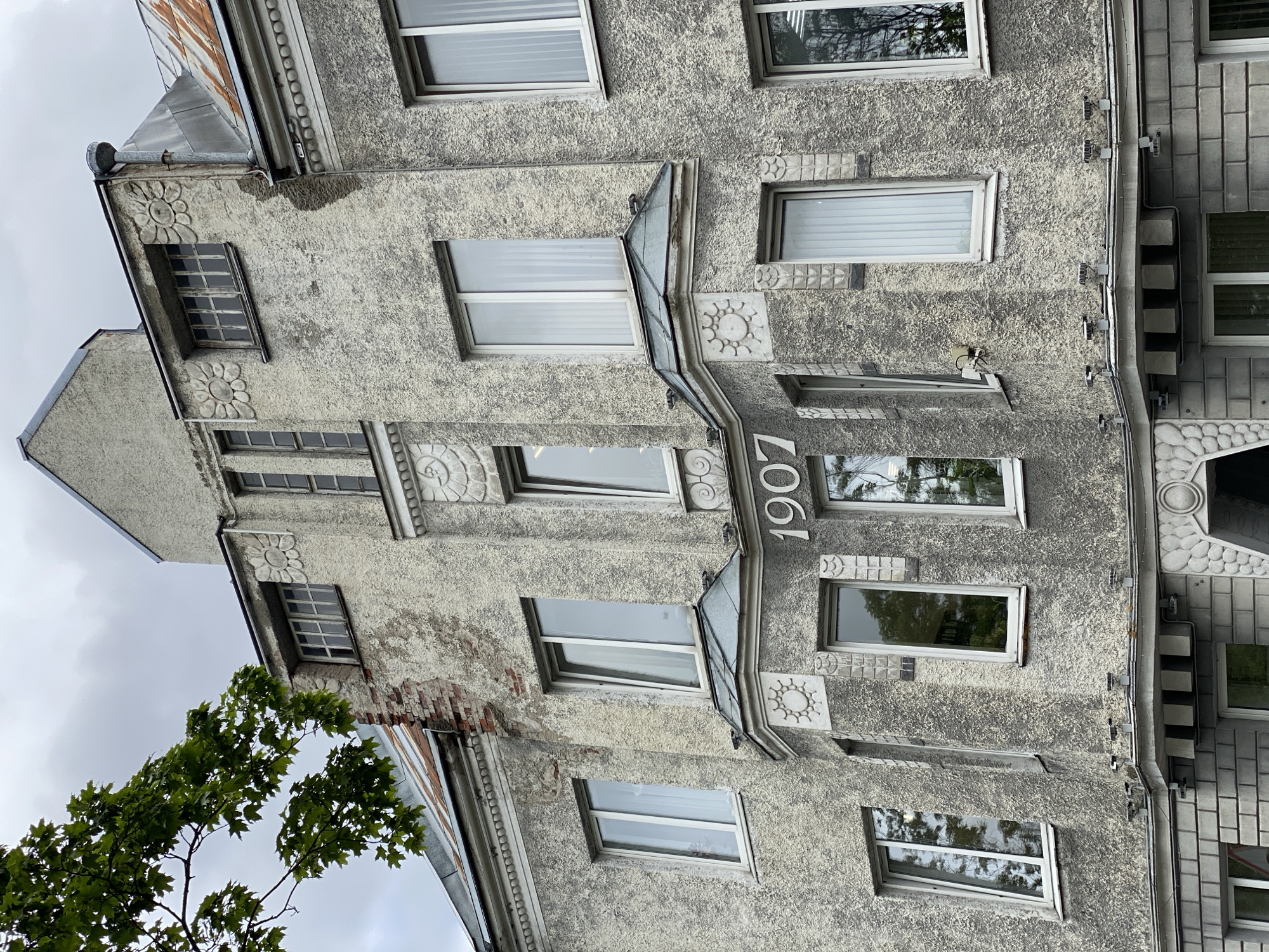 The image shows a close-up, eye-level view of the facade of a historic building in Sortavala, Russia. The building's exterior is a rough, textured grey stucco, showing signs of age and some weathering, with exposed brick in one section. The architectural style appears to be Art Nouveau or a related early 20th-century style, characterized by decorative elements such as floral motifs and molded ornamentation.
Prominently displayed on the central bay of the facade are the white numerals "1907", indicating the year of construction. Numerous windows are visible across different floors, mostly rectangular with white frames and blinds or curtains. Some windows are reflecting the overcast sky and surrounding trees.
The roofline is visible at the top, with some sections appearing to be made of corrugated metal that shows rust. There is a drainpipe running down the side of the building. At the bottom of the image, a portion of the building's base is visible, constructed with grey brickwork and featuring decorative circular motifs. The overall impression is of an aged, architecturally interesting building in an urban setting, with no people or active events depicted.